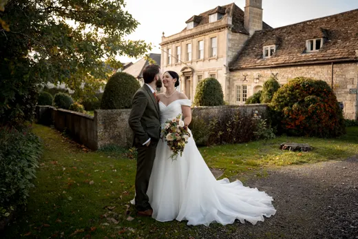 A bride holding an autumn bouquet and a groom in a tweed suit share a tender moment outside a historic country house at golden hour.