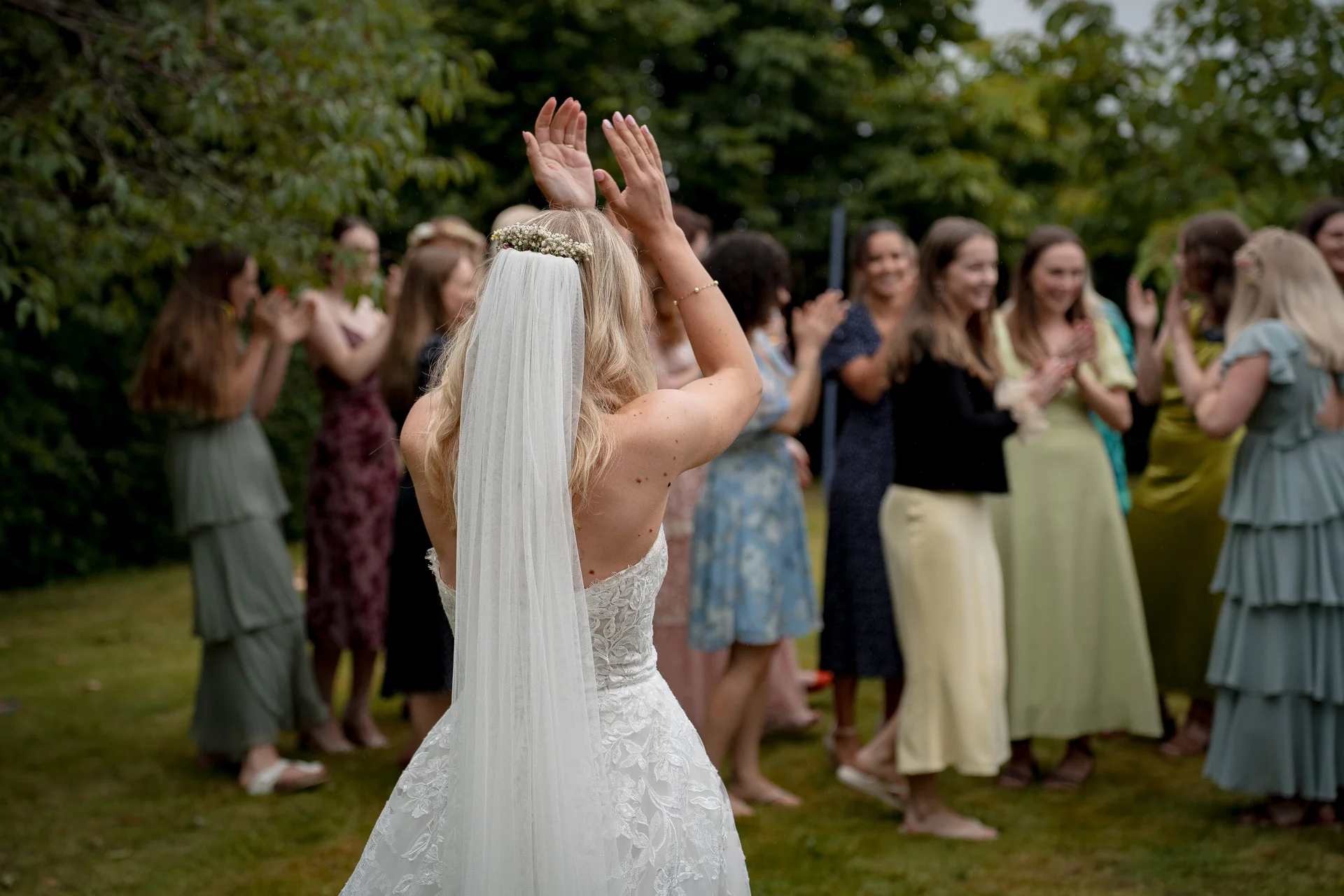 A bride in a lace gown and veil raises her hands in celebration outdoors, surrounded by clapping guests in an array of colourful dresses.