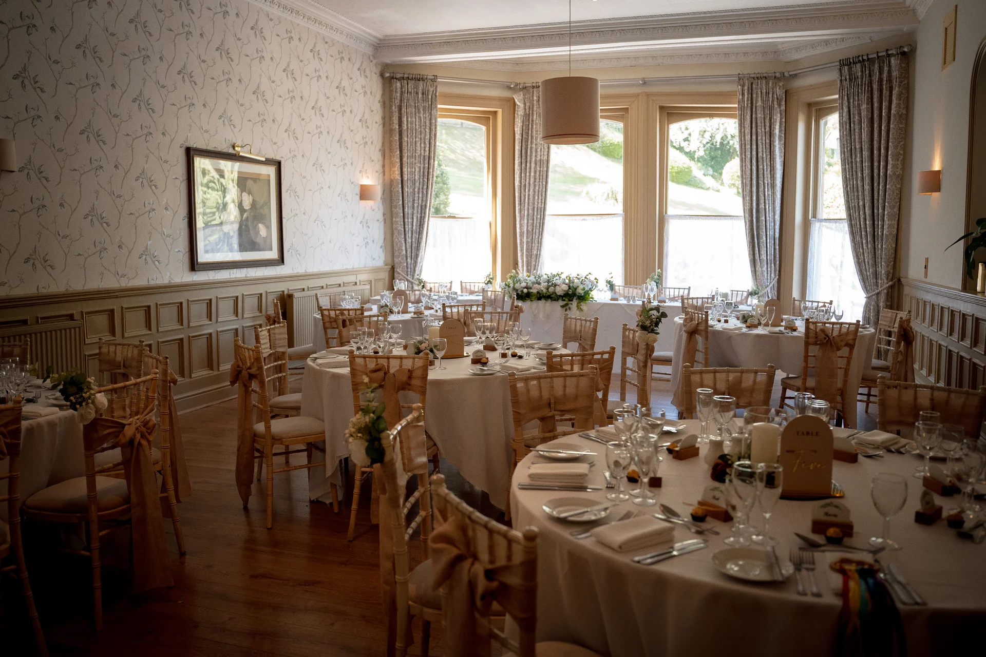 An elegant hotel wedding reception room featuring round tables with white linens, wooden chairs adorned with bows, and floral centrepieces under natural window light.