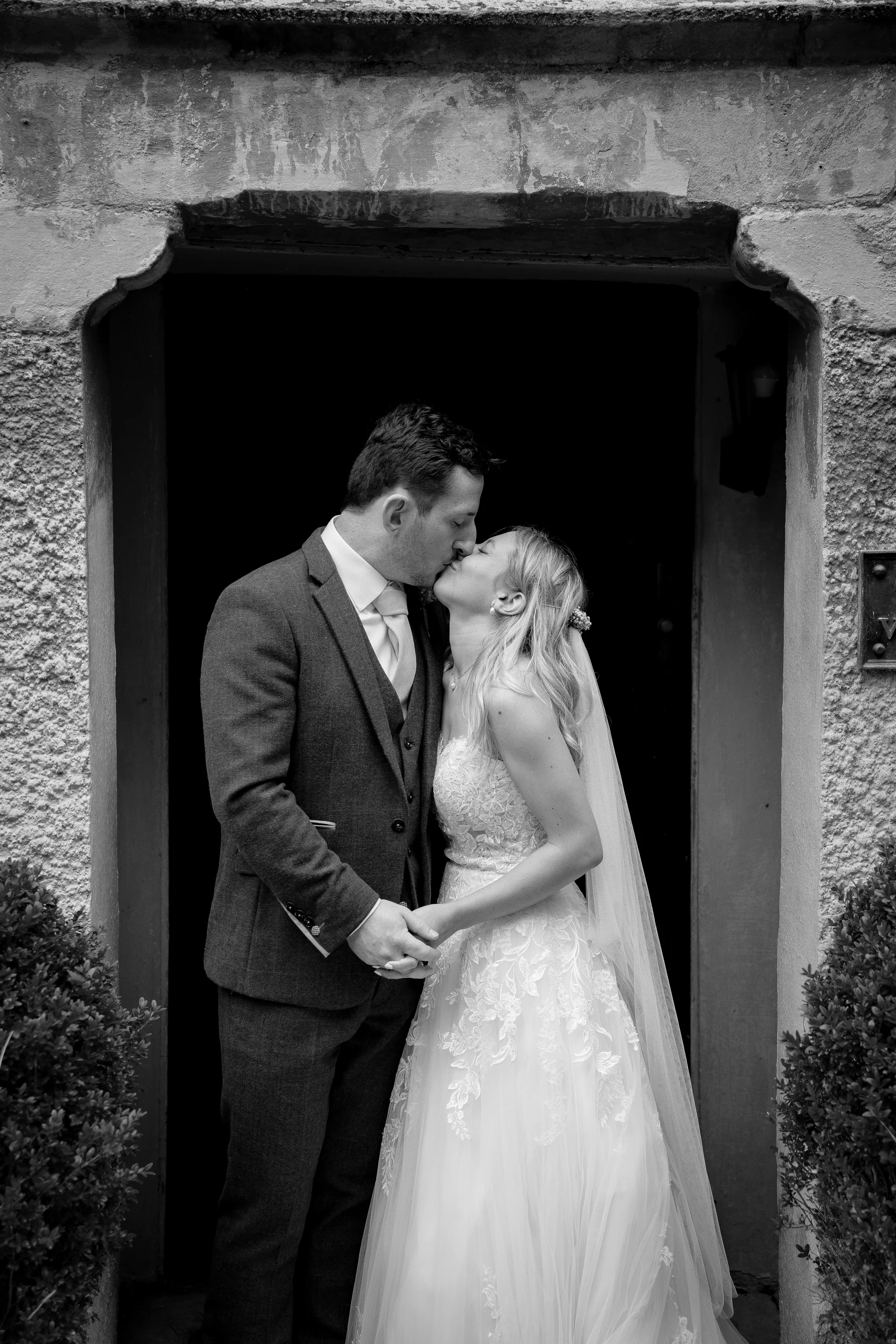 Newlyweds share a tender kiss while standing hand in hand inside a stone doorway, captured in a timeless black and white portrait.