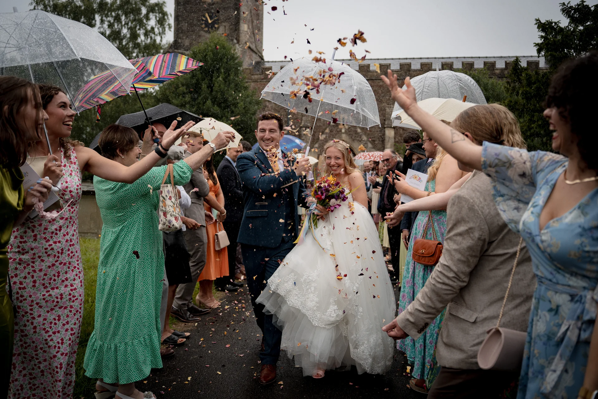 A newlywed couple joyfully walks through a shower of colourful confetti outside a historic church while guests hold up transparent umbrellas in the rain.