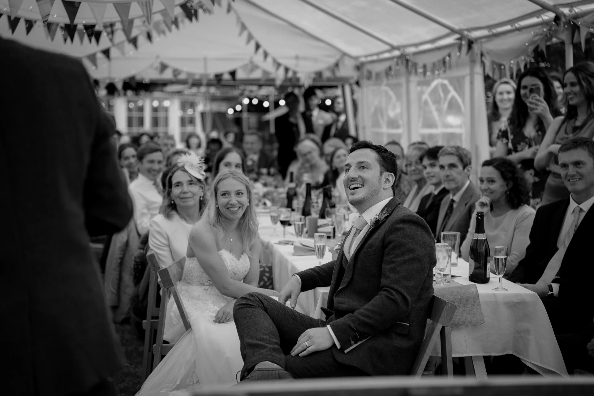 A newlywed couple seated at a top table in a marquee laugh joyfully while listening to wedding speeches, surrounded by smiling guests and festive bunting.