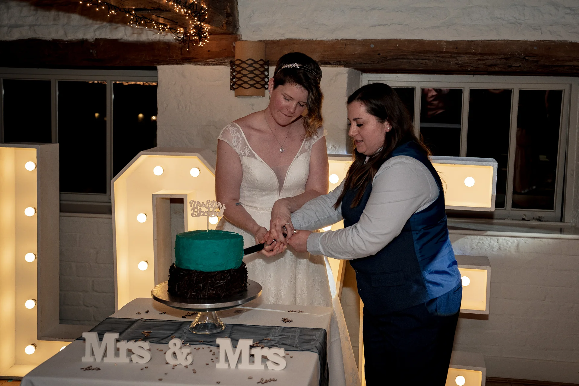 Two brides smiling as they cut a two-tier teal and chocolate wedding cake in front of illuminated marquee letters inside a rustic barn.