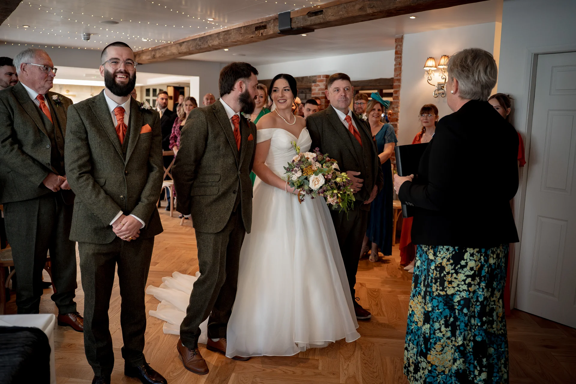 The bride and groom stand smiling at the altar during a civil ceremony, flanked by groomsmen in green tweed suits with burnt orange ties while guests watch on.