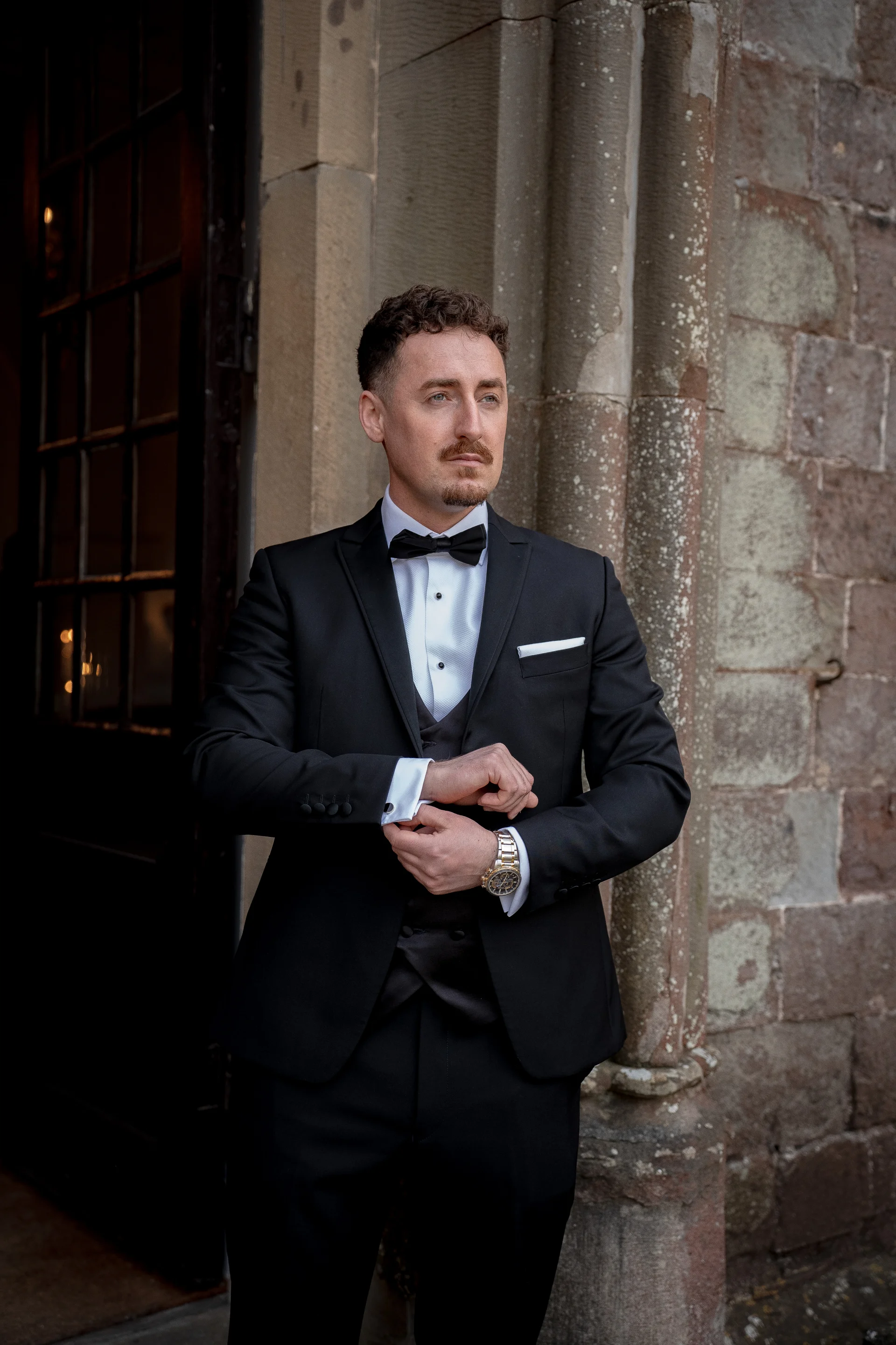 A handsome groom in a sharp black tuxedo adjusts his cuffs while standing outside a historic stone building.