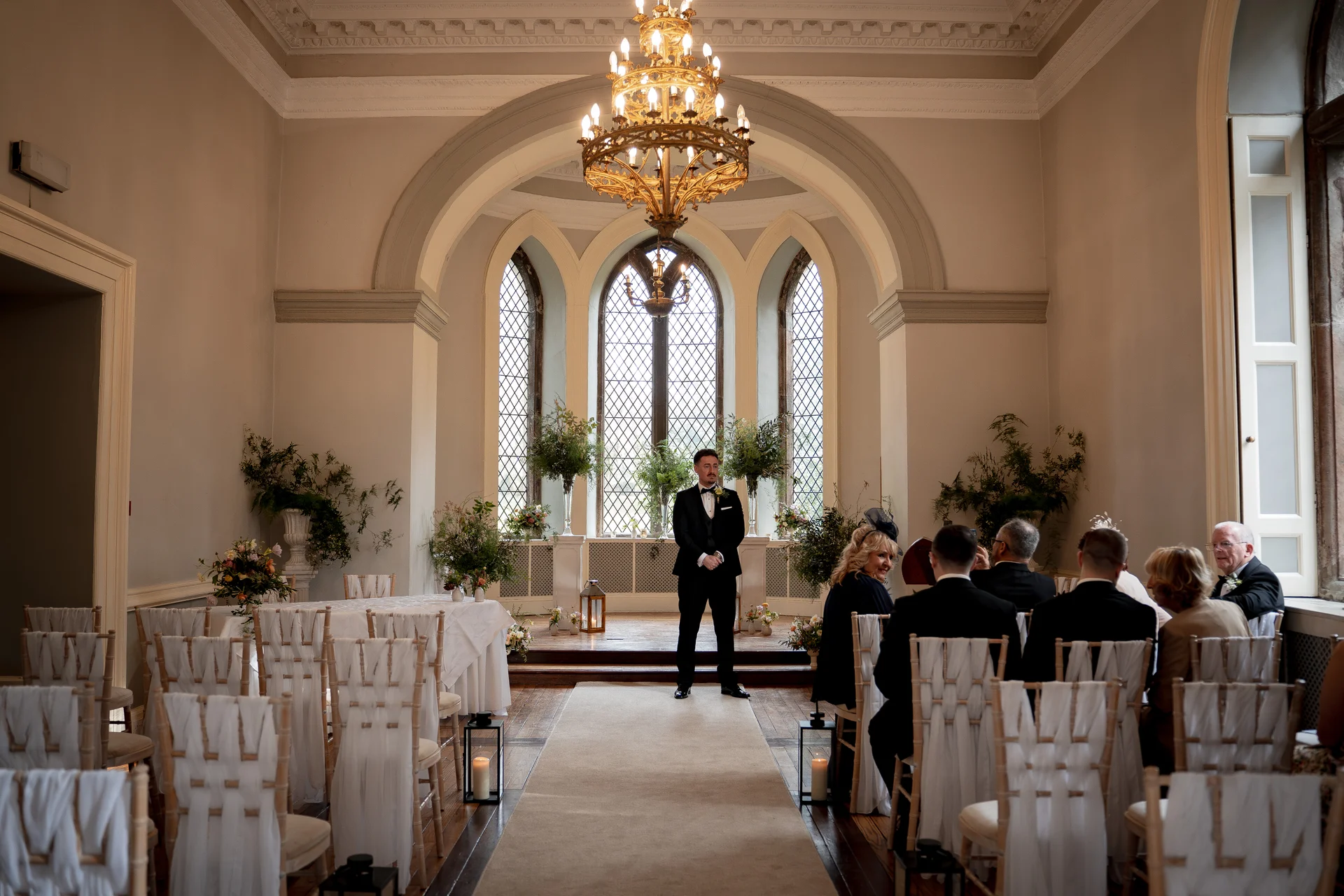 A groom in a classic black tuxedo waits at the front of a grand ceremony room with tall arched windows and a large chandelier.