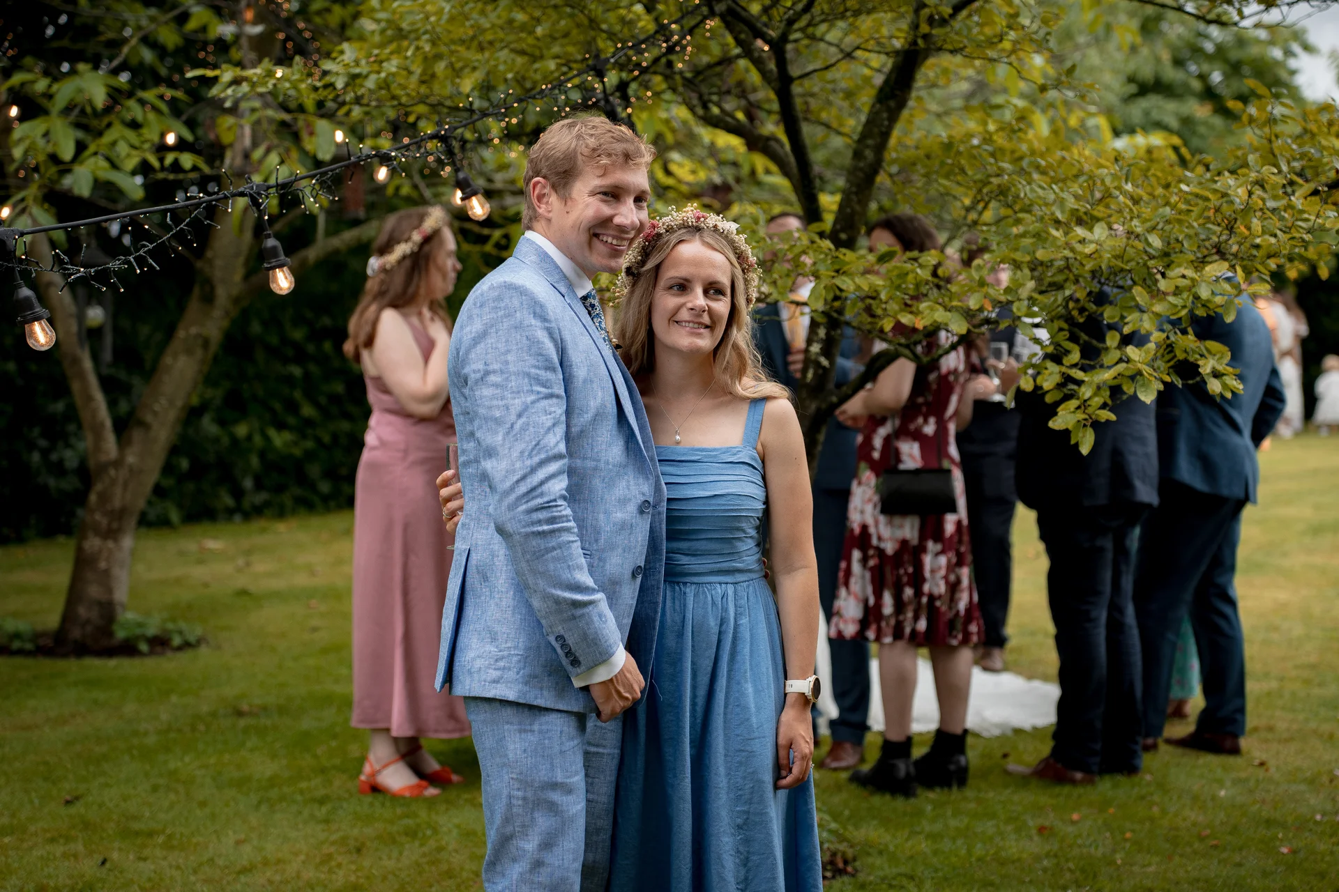 A smiling couple embracing outdoors in a garden setting, with the groom wearing a light blue linen suit and the bride in a blue dress with a floral crown.