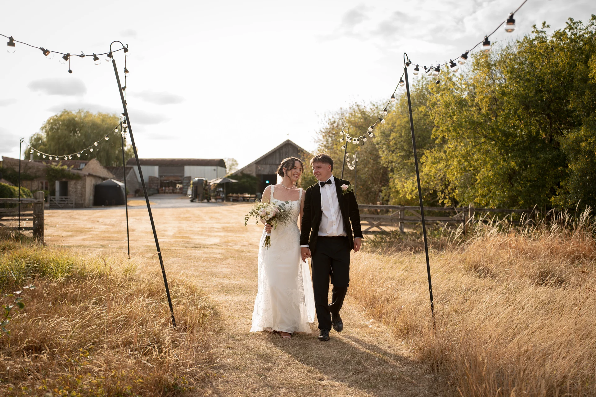 A smiling bride and groom walk hand in hand through a sunlit field framed by festoon lights, with rustic farm buildings visible in the background.