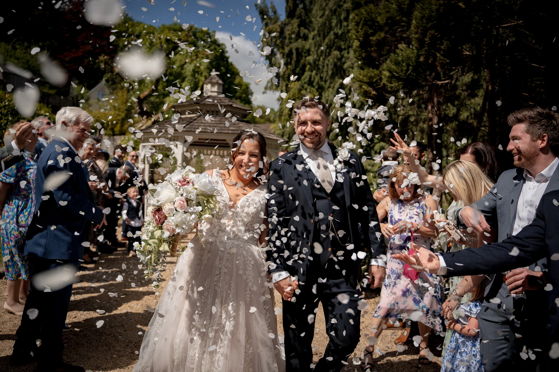 A newlywed couple walks hand in hand through a shower of white confetti as joyful guests cheer them on outdoors.