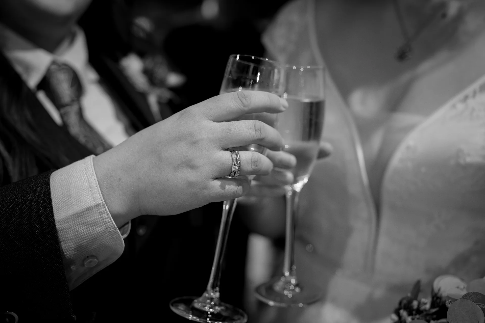 A black and white close-up shot captures a newlywed couple toasting with champagne flutes, showcasing their wedding bands and engagement ring.