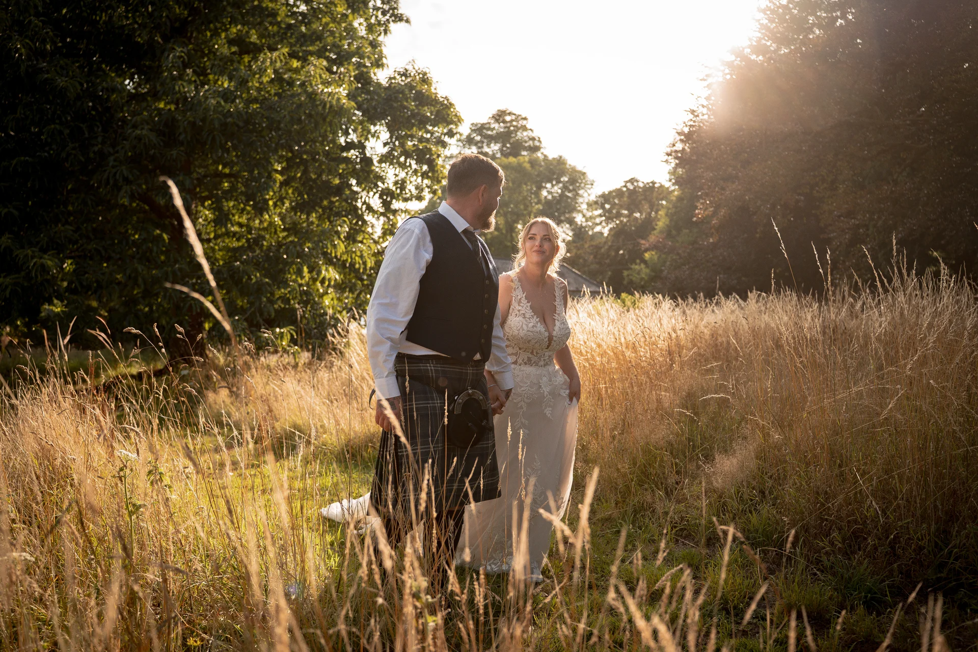 A newlywed couple walks hand in hand through a sunlit tall grass meadow during golden hour, with the groom wearing a traditional kilt and the bride in an embroidered lace gown.