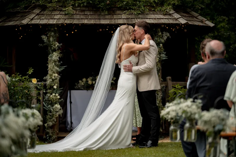 A newly married bride and groom share their first kiss in front of a flower-adorned wooden gazebo while guests watch from the aisle.