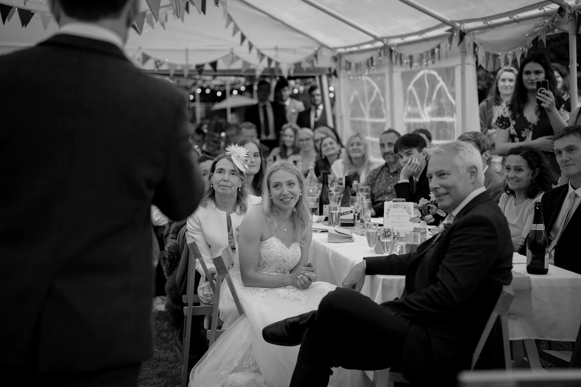 A black and white documentary photograph captures guests seated at a marquee wedding breakfast while listening attentively to a speech.