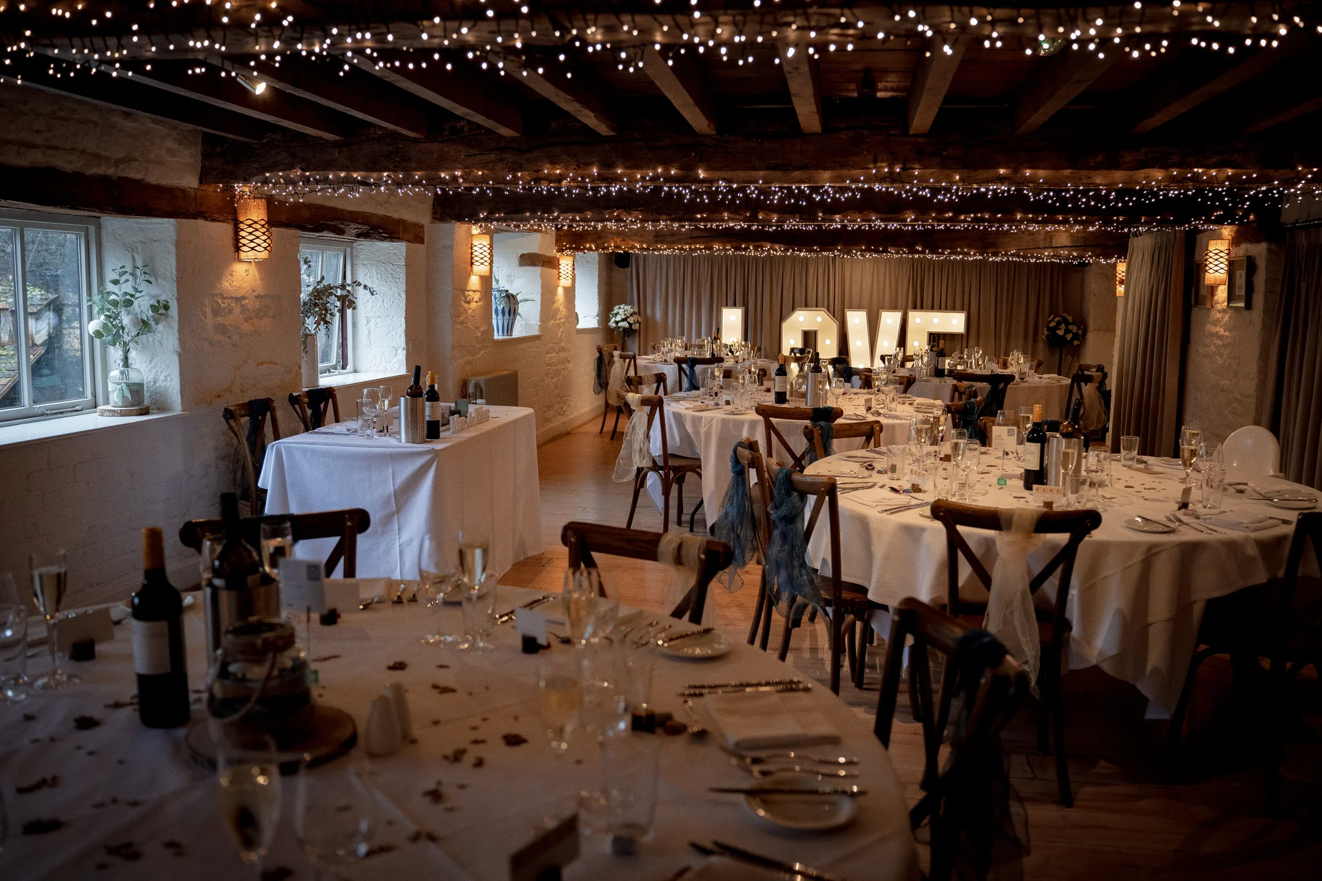 An atmospheric interior view of a rustic barn wedding reception featuring round tables with white linens, wooden chairs, and a stunning canopy of twinkling fairy lights across the exposed ceiling beams.