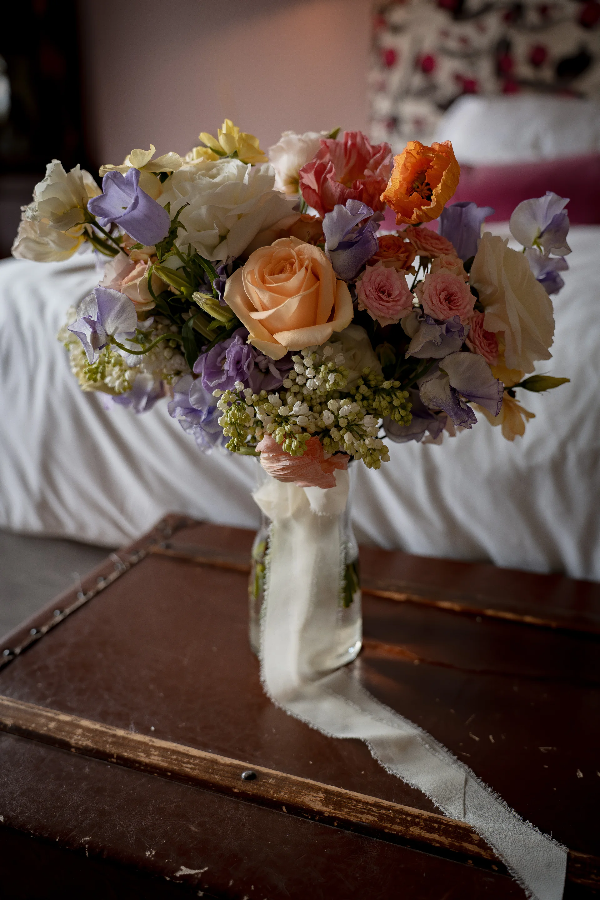 A colourful garden style wedding bouquet featuring peach roses, purple sweet peas, and ranunculus sits in a clear glass vase with a long trailing silk ribbon on a dark wooden surface.