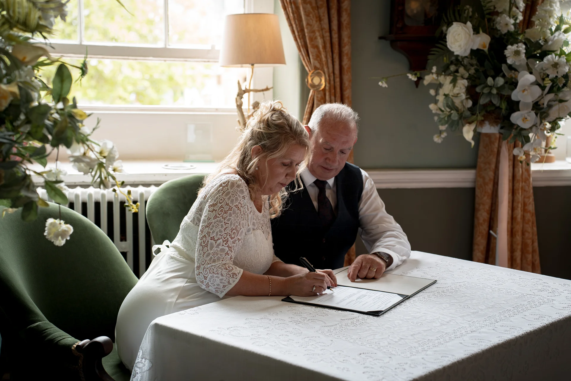 An older couple signs the wedding register together at a table decorated with a white lace cloth inside a bright room.