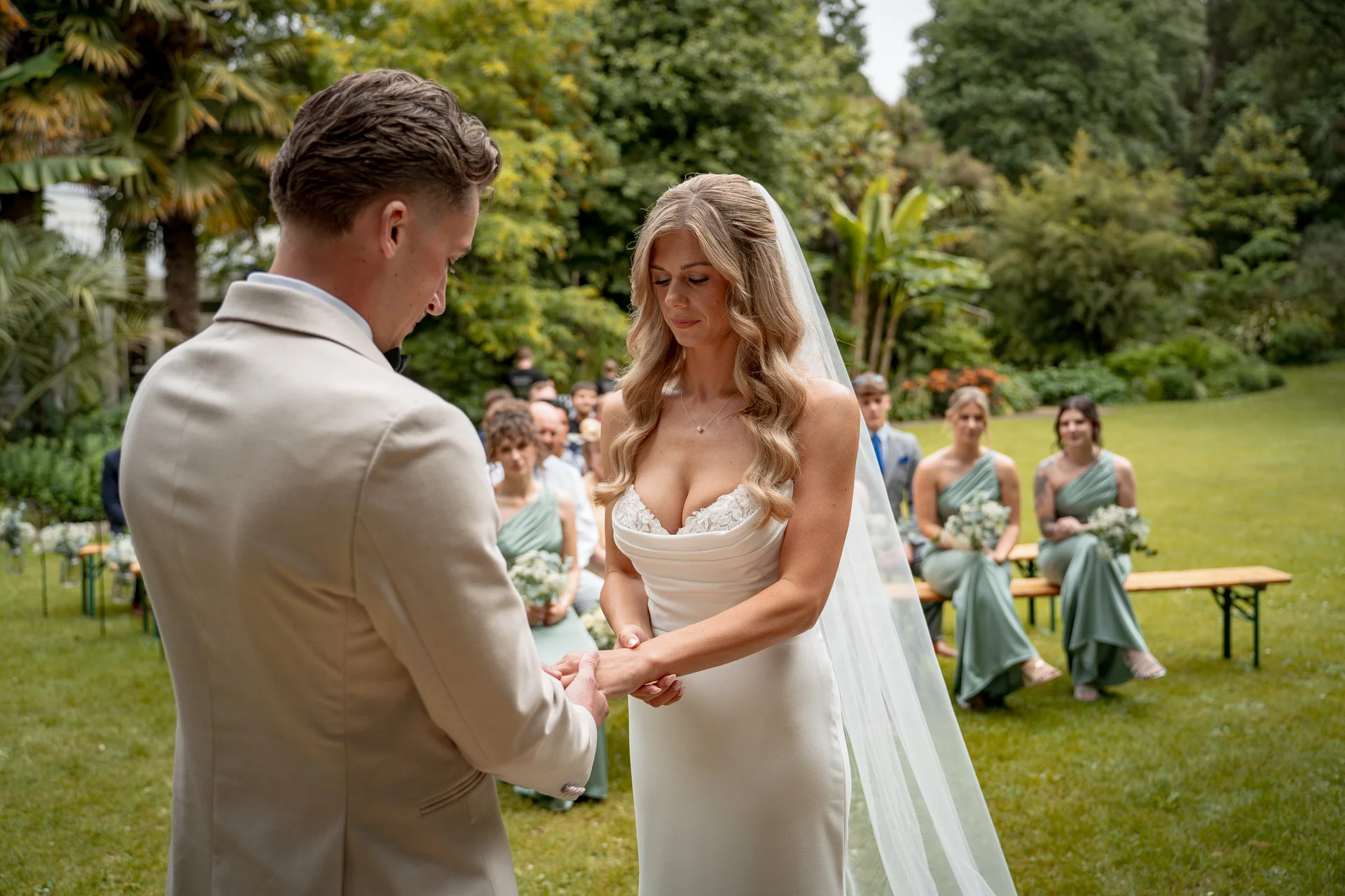 A groom in a beige suit places a ring on the bride's finger during an outdoor garden ceremony, with bridesmaids seated on wooden benches in the background.