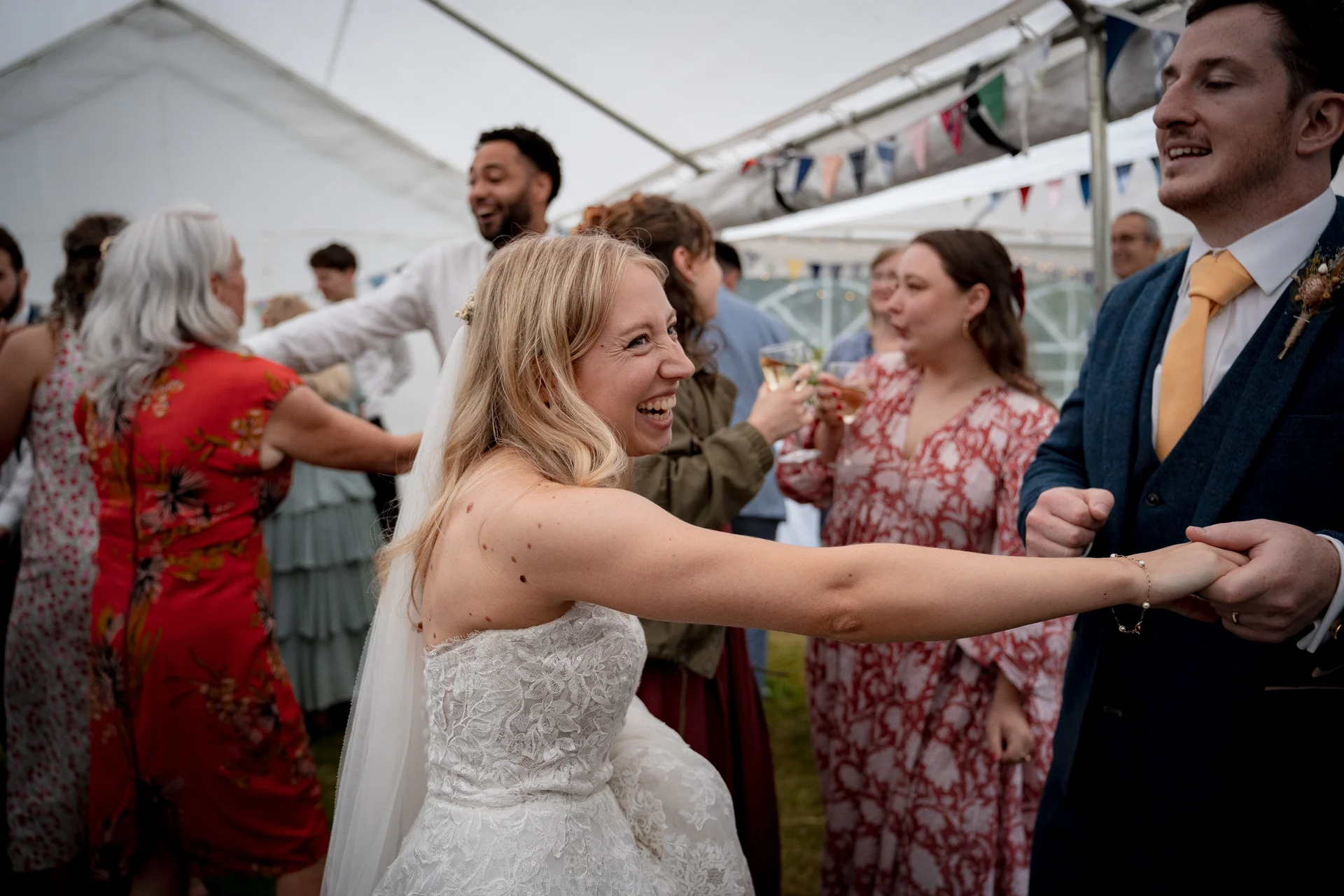 A joyful bride and groom laugh while dancing together inside a marquee decorated with bunting, surrounded by celebrating wedding guests.