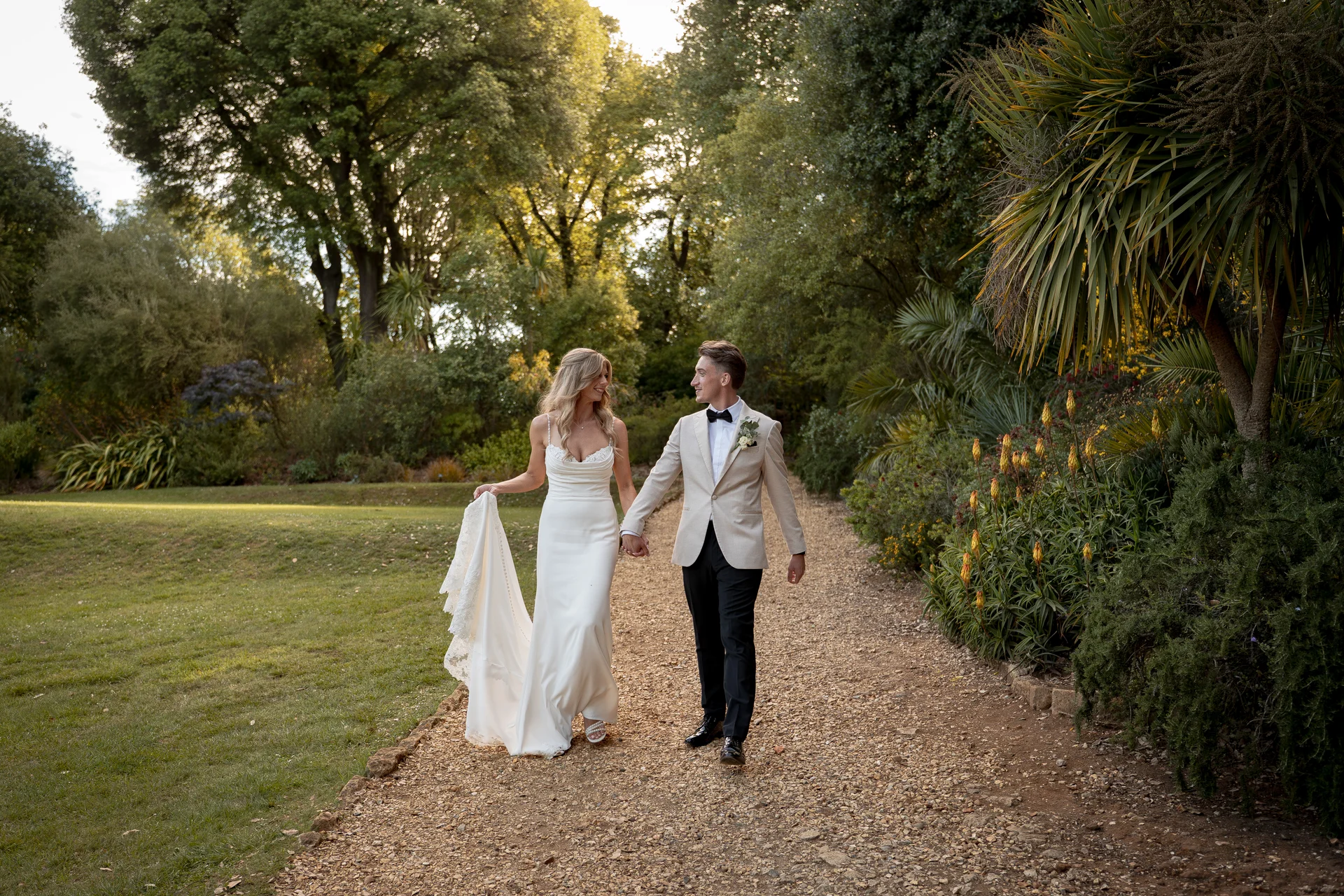 A bride and groom walk hand in hand along a sunlit gravel path through lush gardens during the golden hour, smiling affectionately at each other.