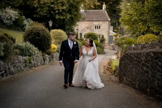 A newlywed couple holds hands and smiles at each other while walking down a scenic country lane beside a stone wall and historic building.