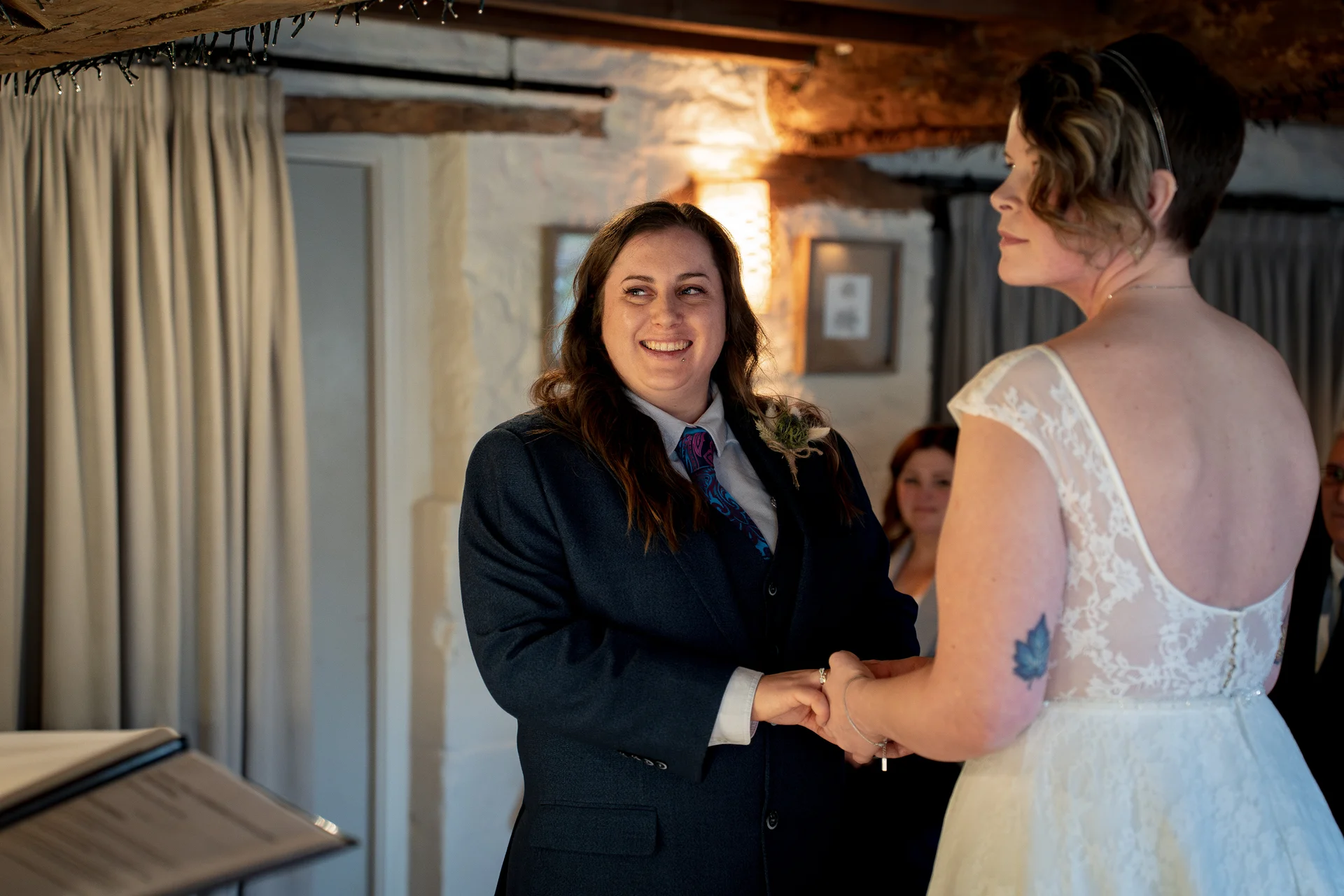 Two brides stand holding hands and smiling during their indoor wedding ceremony inside a rustic barn with exposed wooden beams.