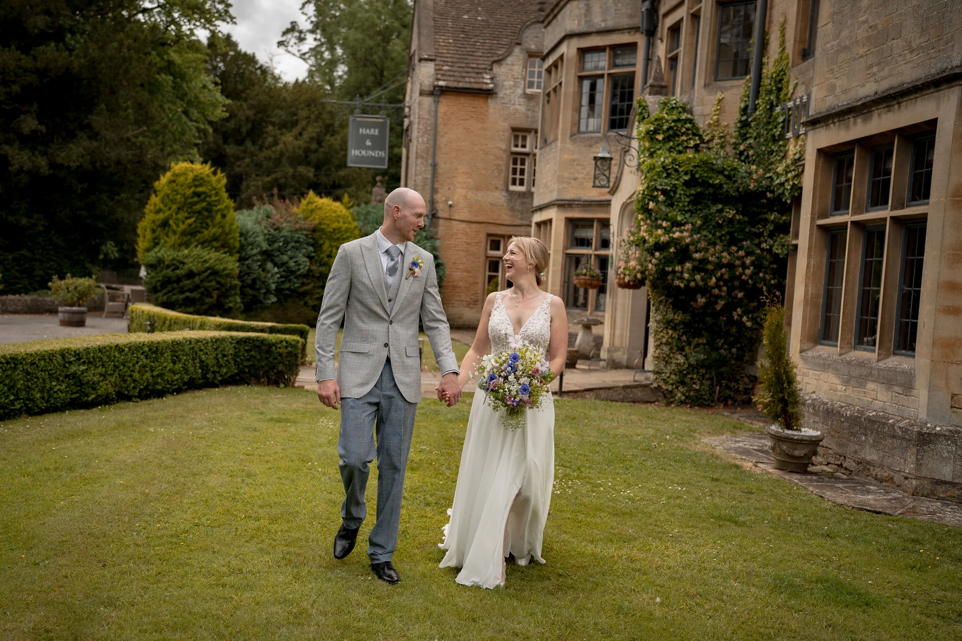 A smiling bride and groom walk hand in hand across the lawn of a historic stone country house while looking at each other.