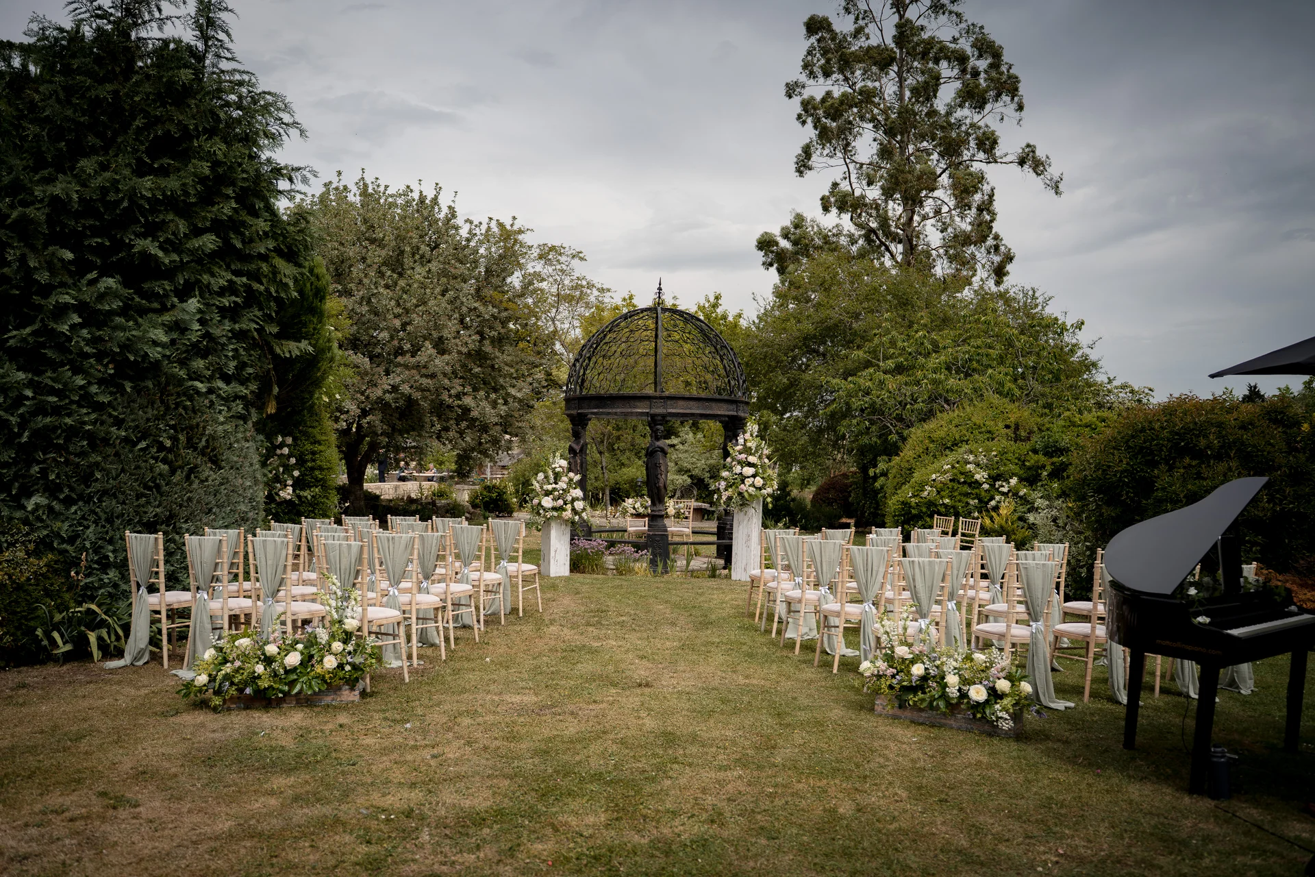 An outdoor wedding ceremony setup features rows of chairs draped in sage green fabric leading towards a dark metal gazebo adorned with floral arrangements, with a grand piano placed to the side on a grassy lawn under an overcast sky.
