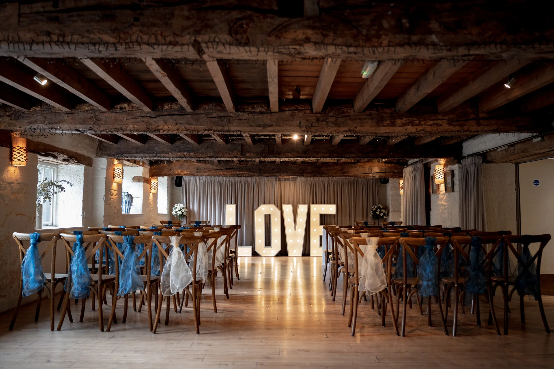 A rustic barn wedding ceremony venue features rows of wooden chairs adorned with blue and white sashes, facing an illuminated love sign against a draped backdrop.