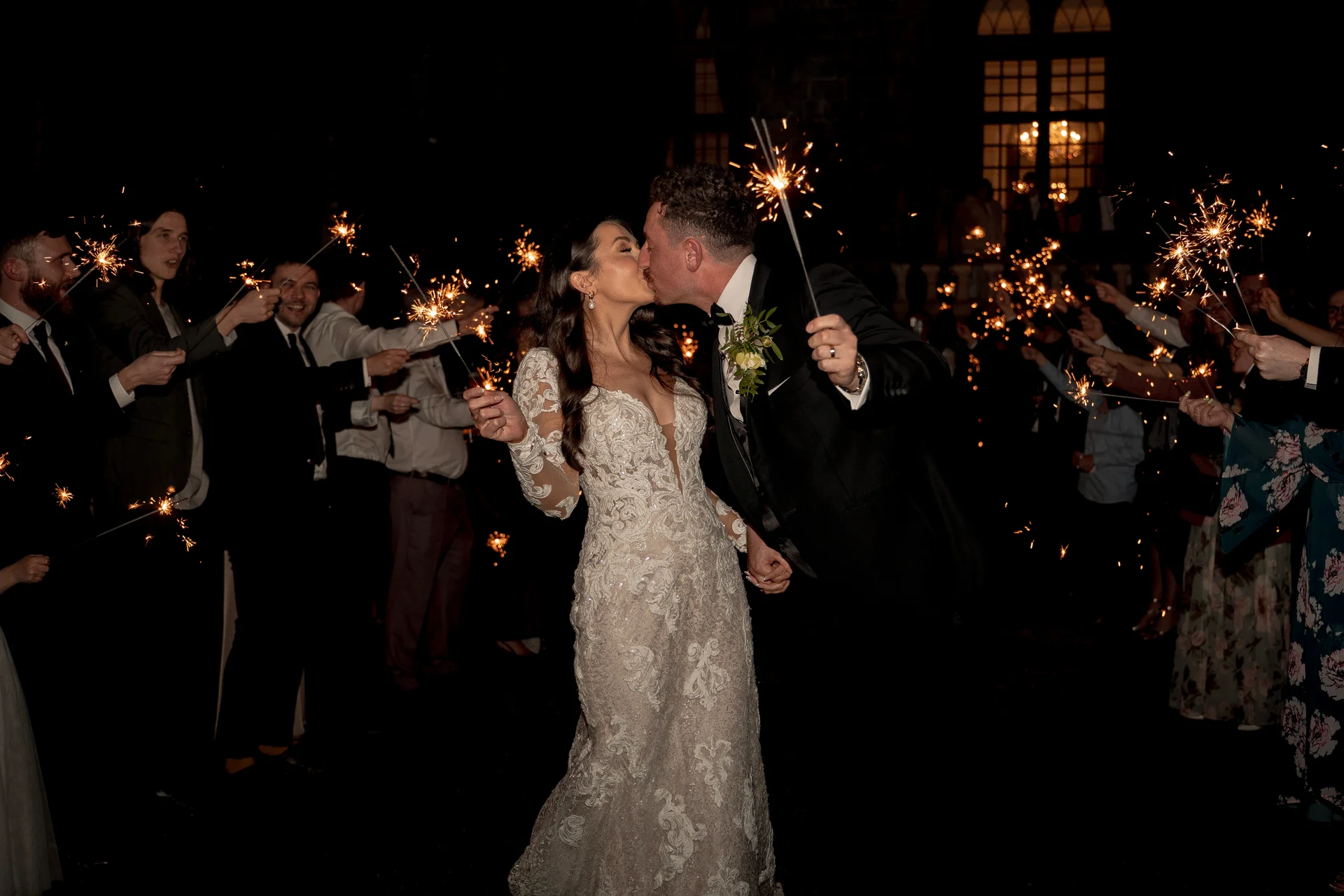 A newlywed couple shares a tender kiss under a sparkling tunnel of handheld sparklers surrounded by cheering wedding guests at night.