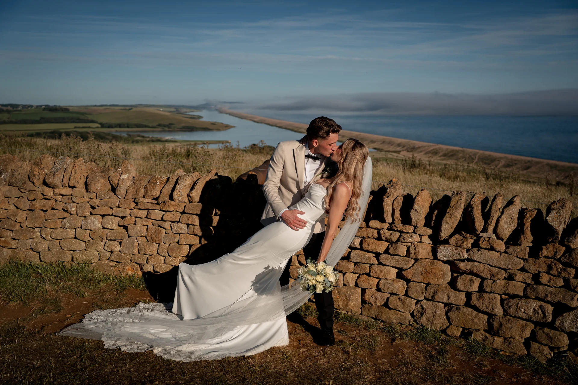 A newlywed couple share a dramatic dip kiss against a stunning coastal backdrop with a stone wall during golden hour.