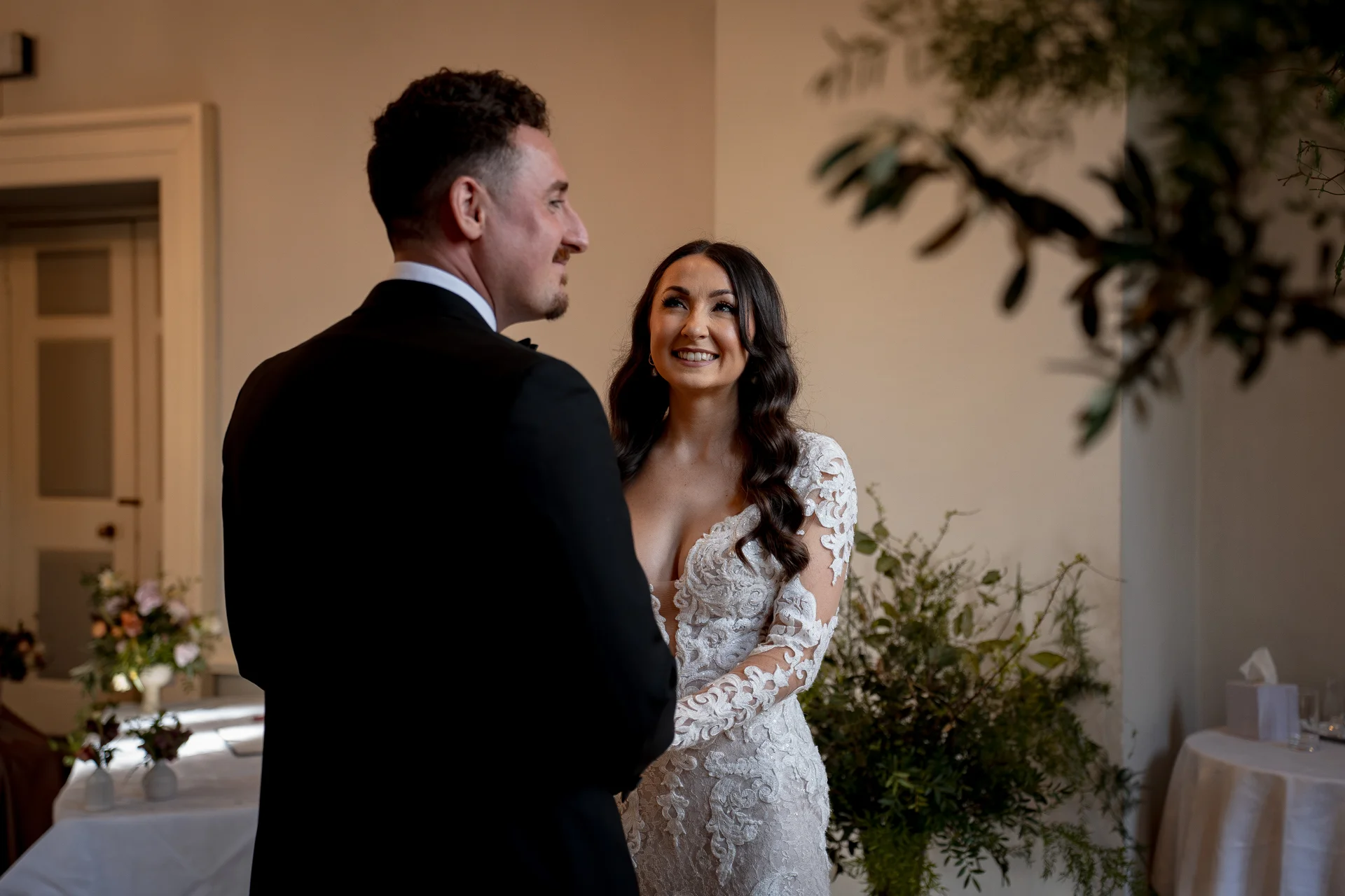 A smiling bride in a lace wedding gown gazes affectionately at her groom in a classic black tuxedo during their ceremony.