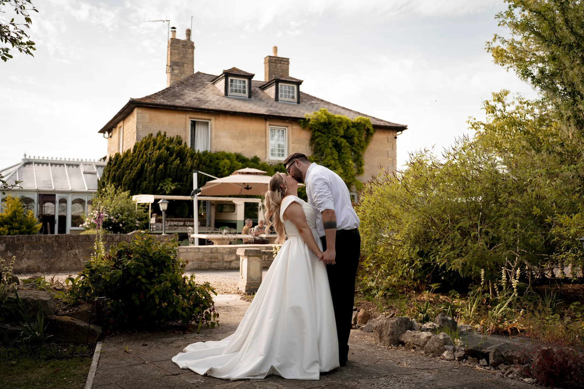 A bride in an a-line satin gown and a groom with rolled sleeves share a tender kiss on a stone patio in front of a historic country house at golden hour.