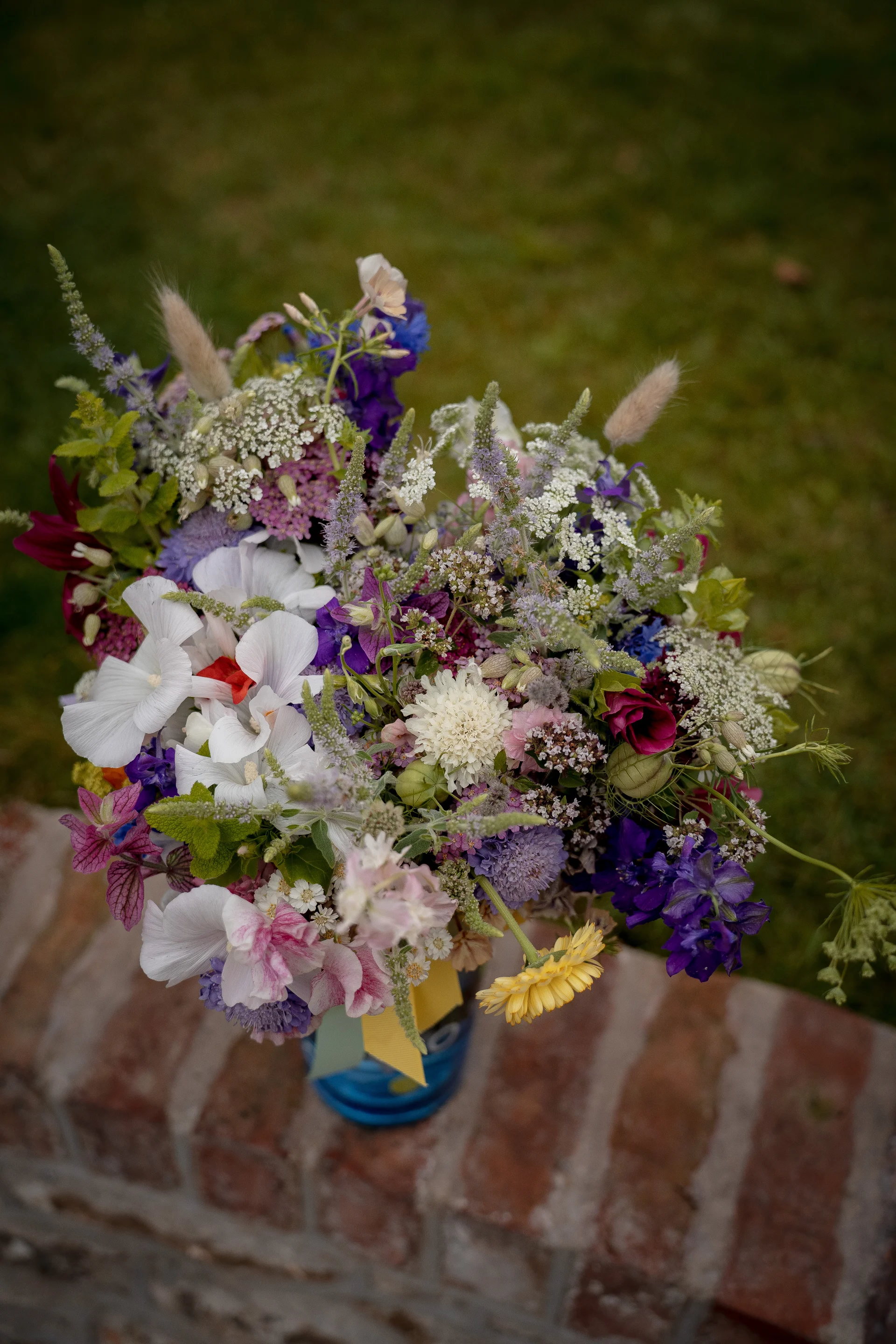 An overhead close-up shot of a vibrant, wild-style wedding bouquet arranged in a blue glass vase resting on a brick ledge.