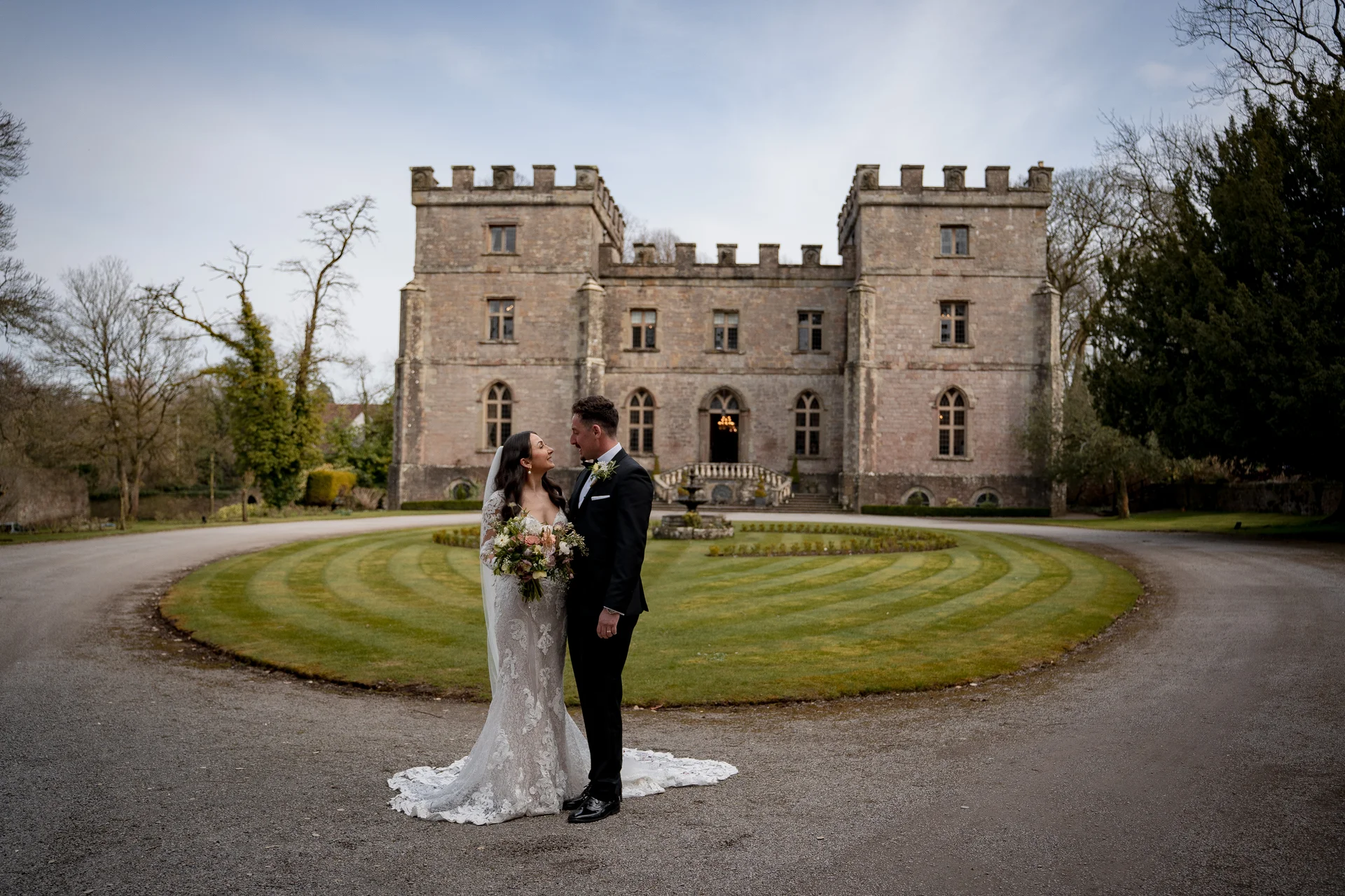 A newlywed couple gazes lovingly at each other on the circular driveway in front of a grand stone castle wedding venue.