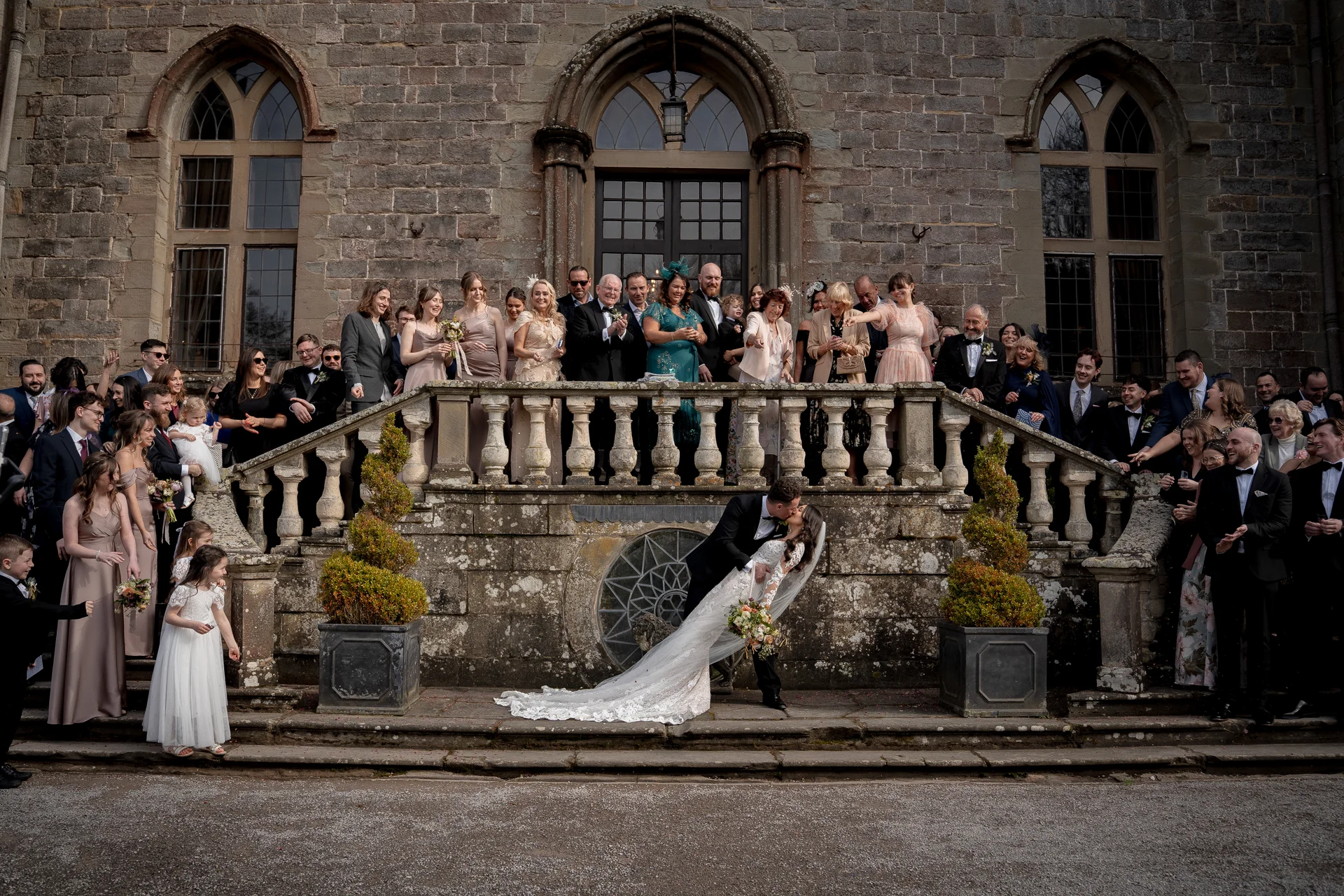 A newlywed couple shares a dramatic dip kiss at the foot of a grand stone staircase outside a historic castle venue, surrounded by cheering wedding guests.