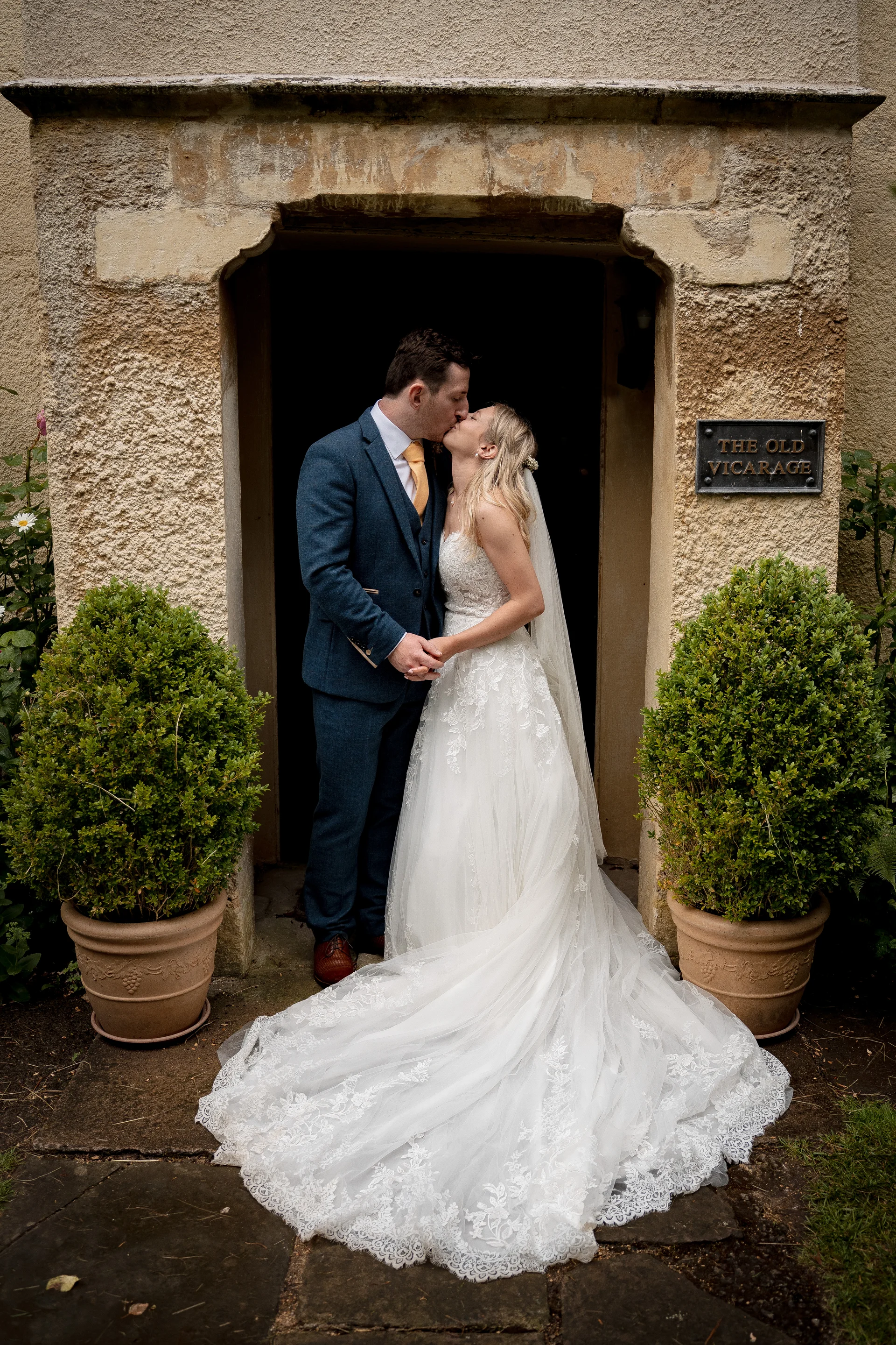 A newlywed couple shares a tender kiss framed by the stone doorway of a historic venue flanked by potted topiary plants.