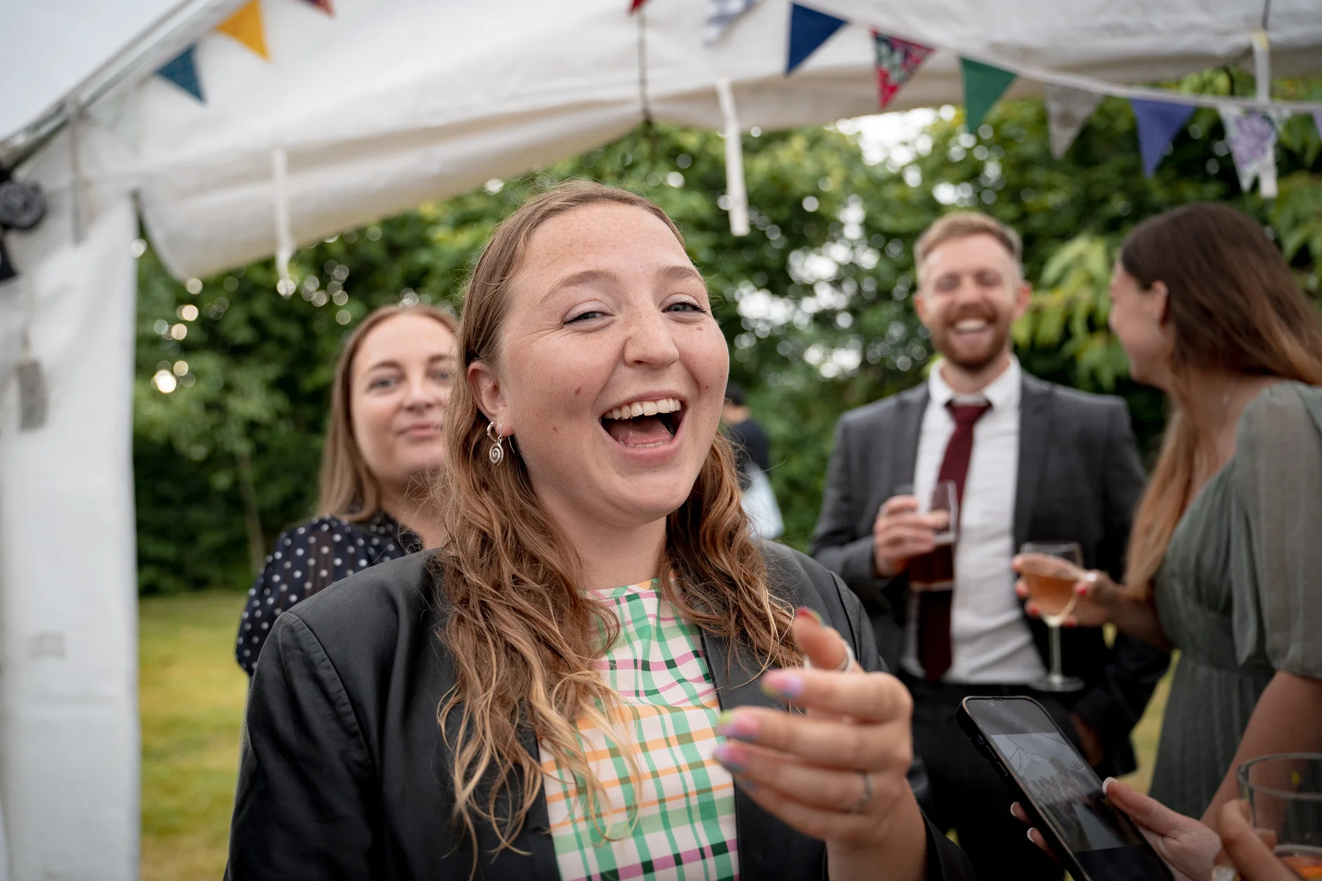 A joyful wedding guest laughs heartily amidst a lively outdoor reception scene beneath a marquee draped with colourful bunting.