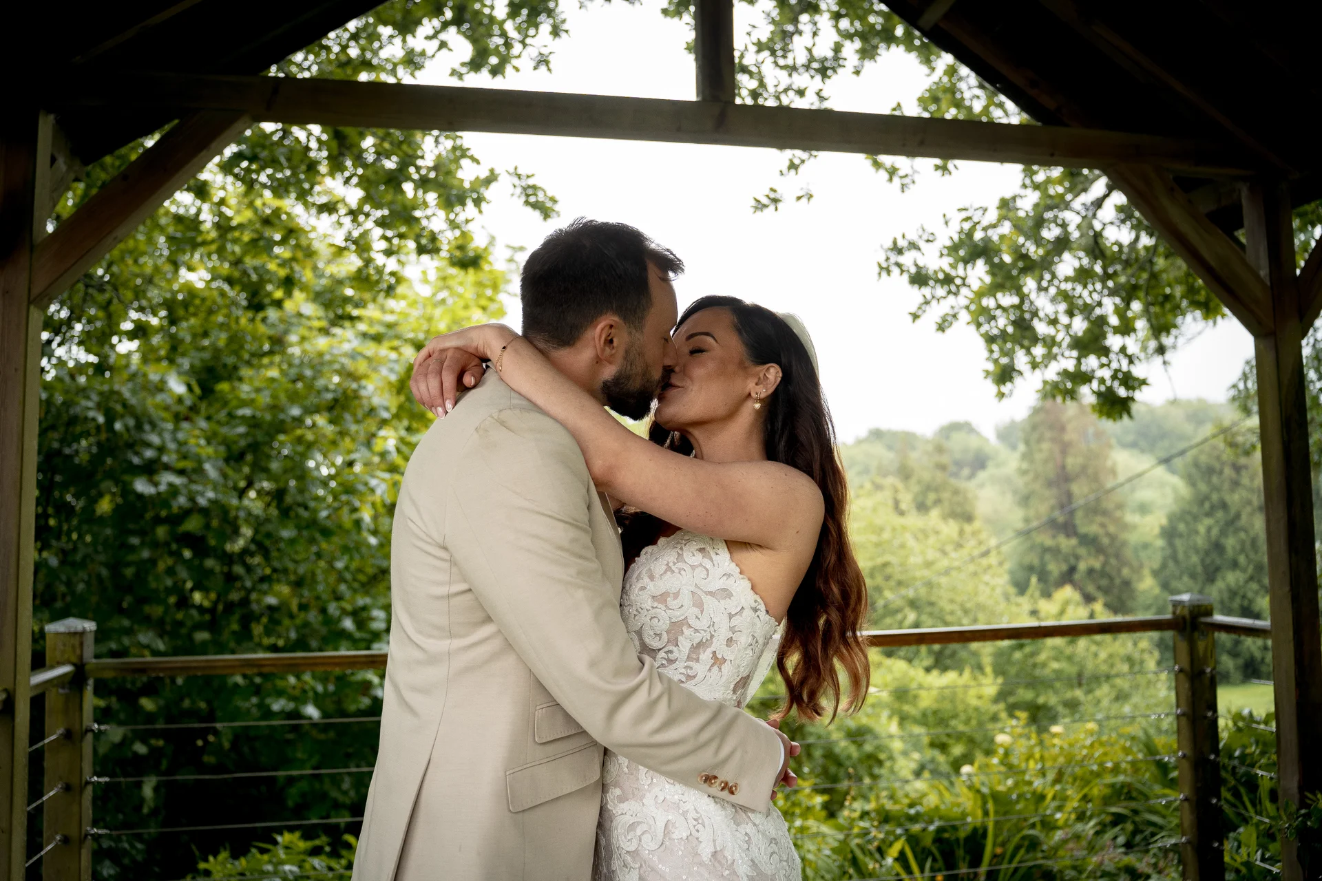 A newlywed couple shares a tender kiss inside a wooden gazebo surrounded by lush green foliage on their wedding day.