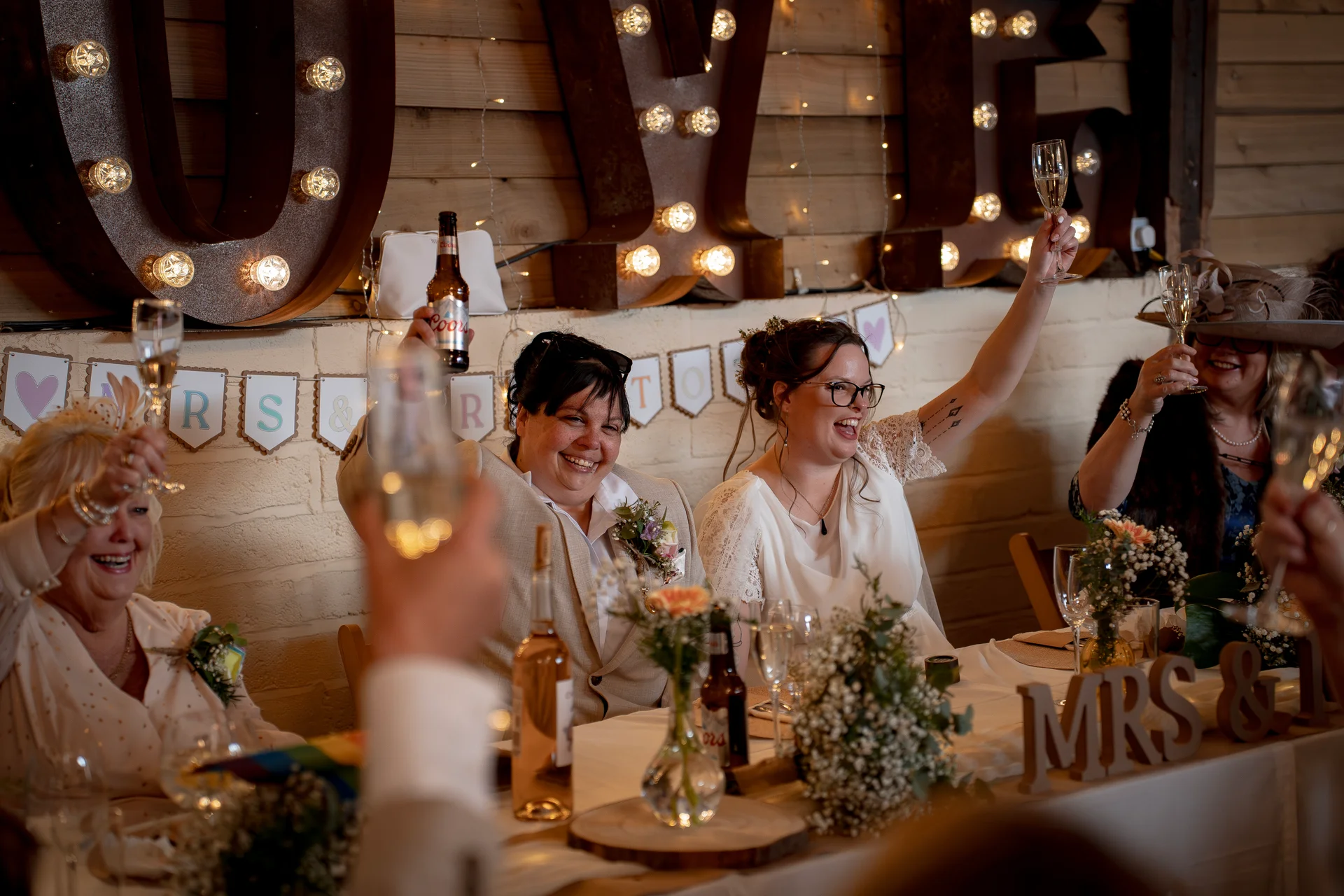 Two brides and guests raise glasses in a joyful toast at the top table during a rustic barn wedding reception.