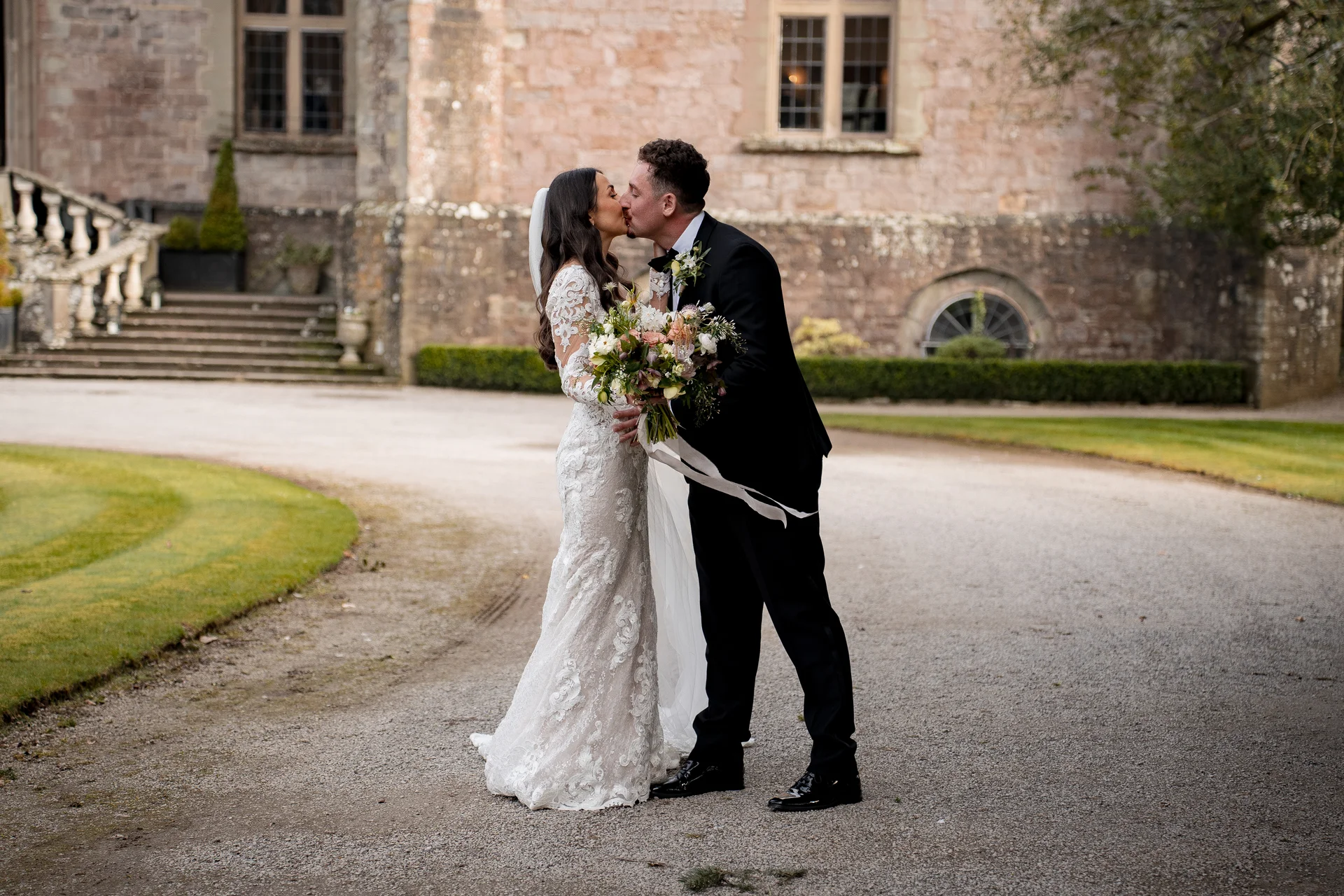 A bride and groom share a tender kiss on a gravel driveway outside a historic stone manor house.