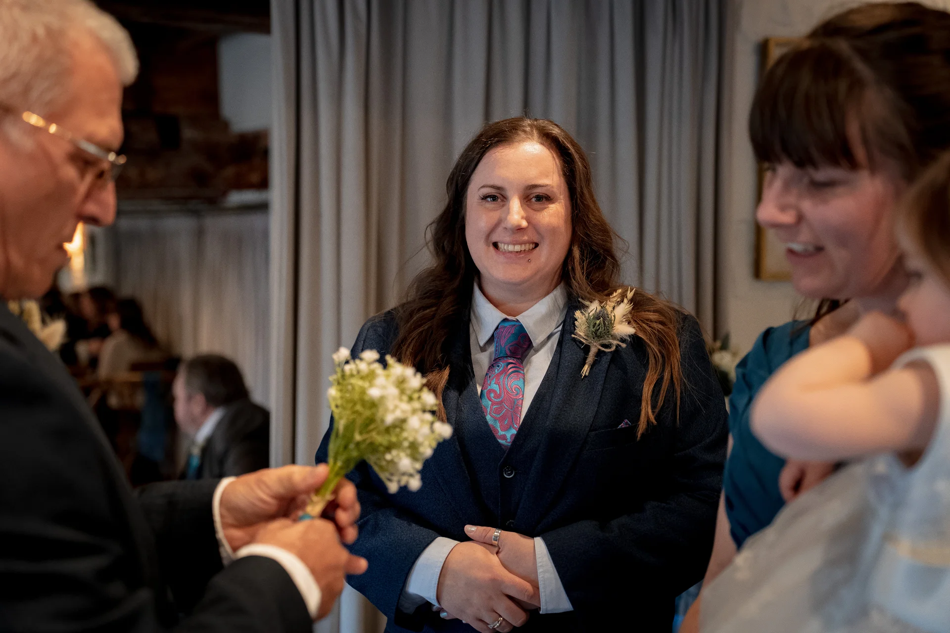 A smiling groom wearing a blue suit and patterned tie holds a small white posy bouquet while surrounded by family and guests.