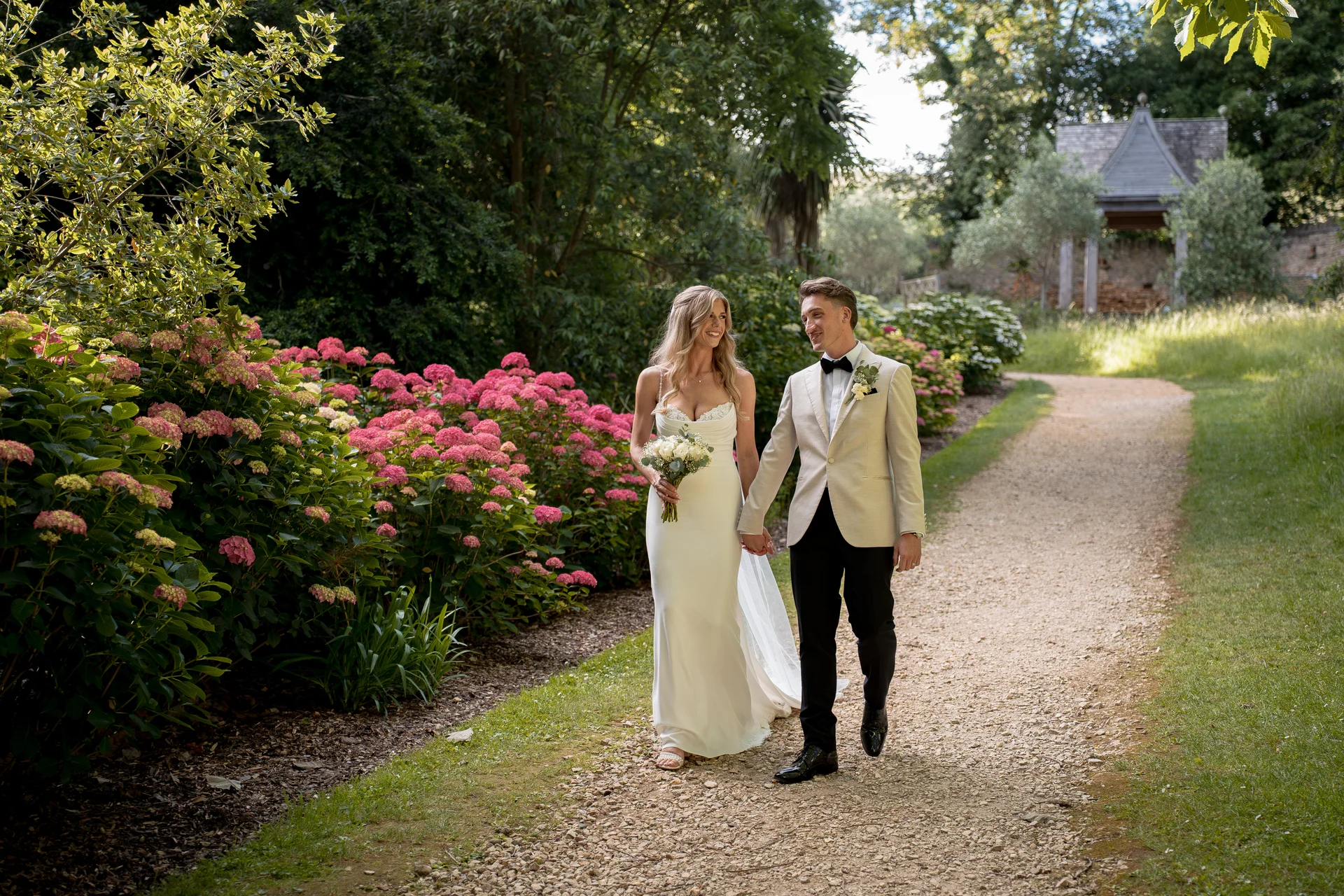 A newlywed couple holds hands and smiles at each other while strolling along a gravel path bordered by vibrant pink hydrangeas and lush green gardens.