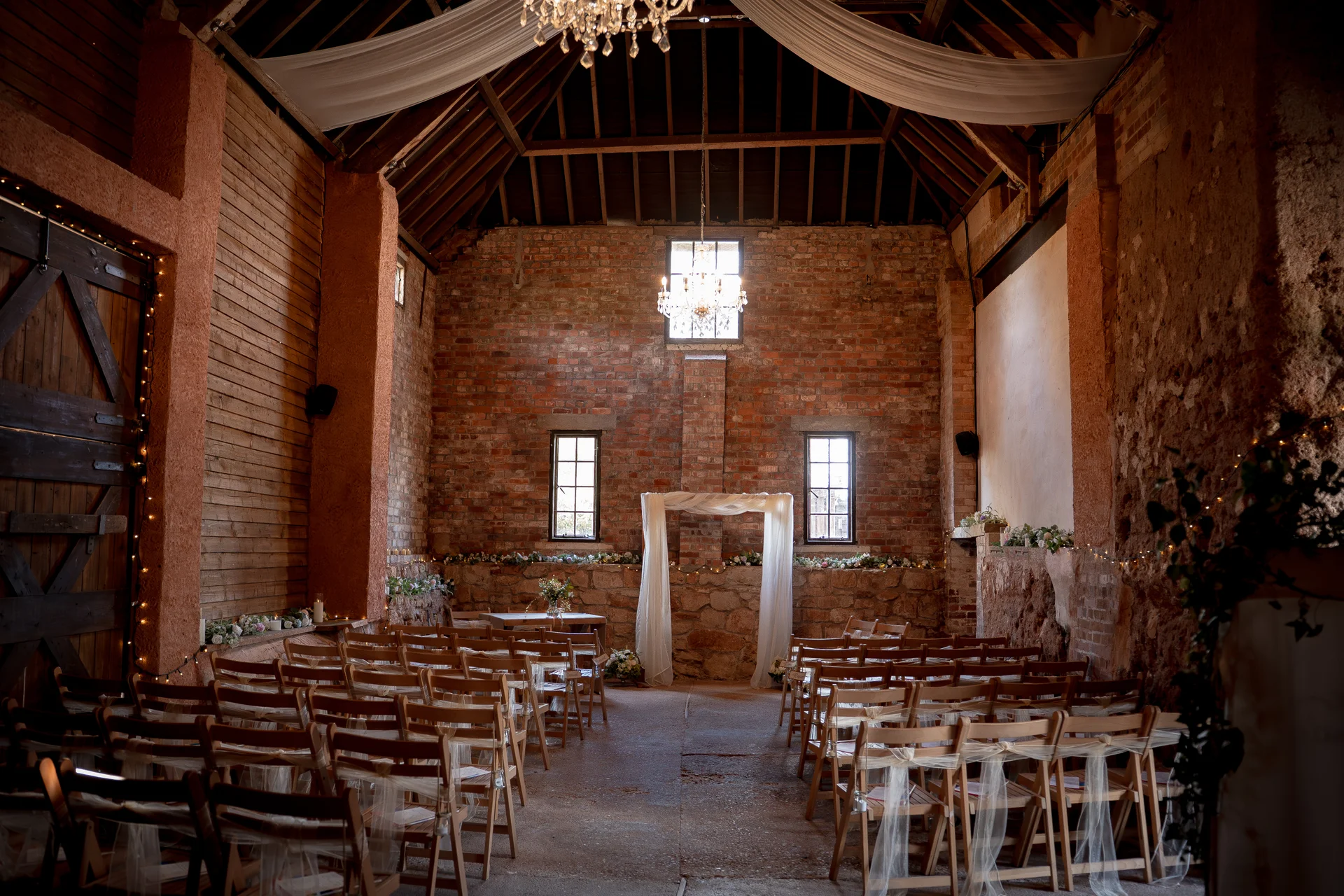 A rustic barn wedding venue featuring exposed brick walls, wooden folding chairs arranged for a ceremony, and a white draped archway illuminated by chandeliers.
