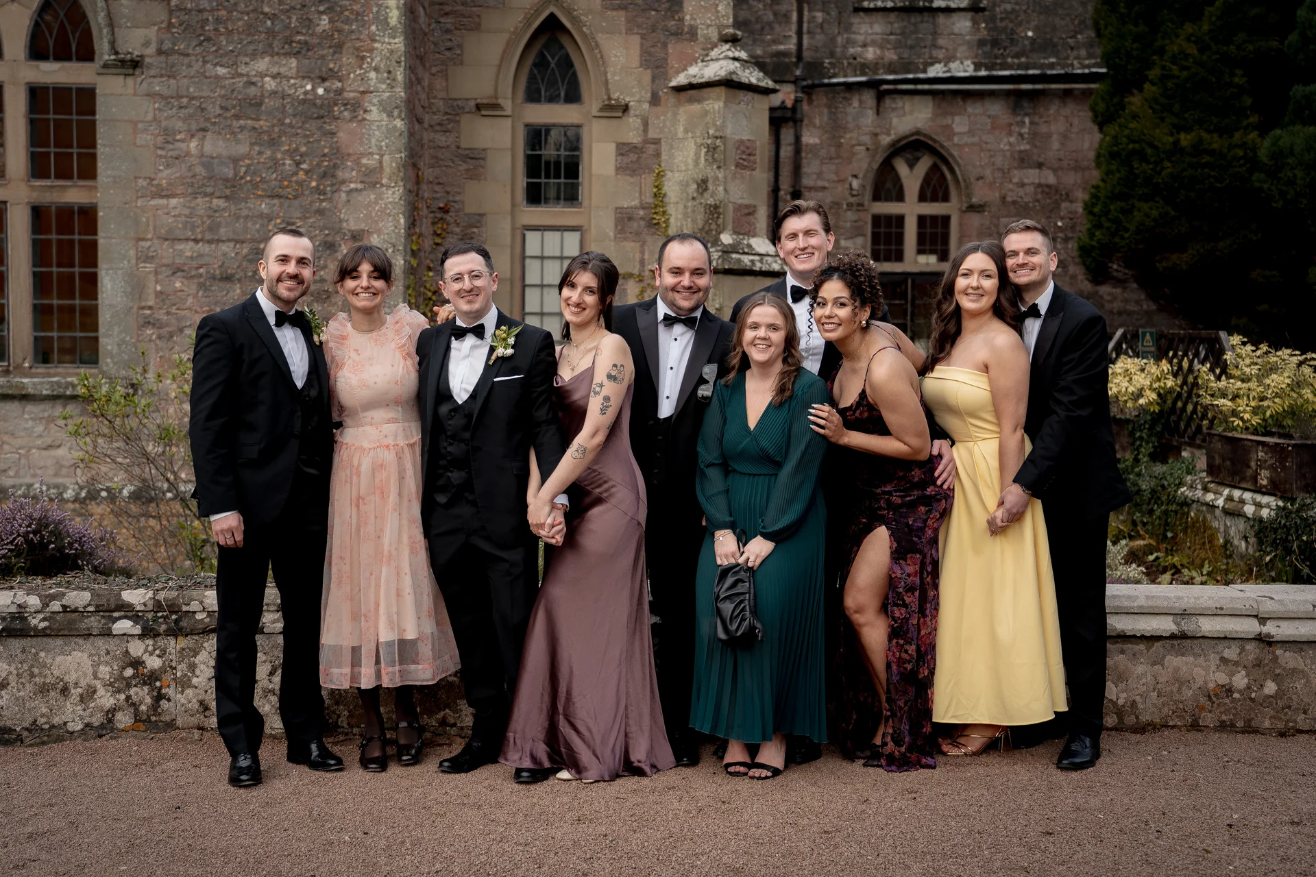 A wedding party and guests pose together outdoors in front of a historic stone castle facade.