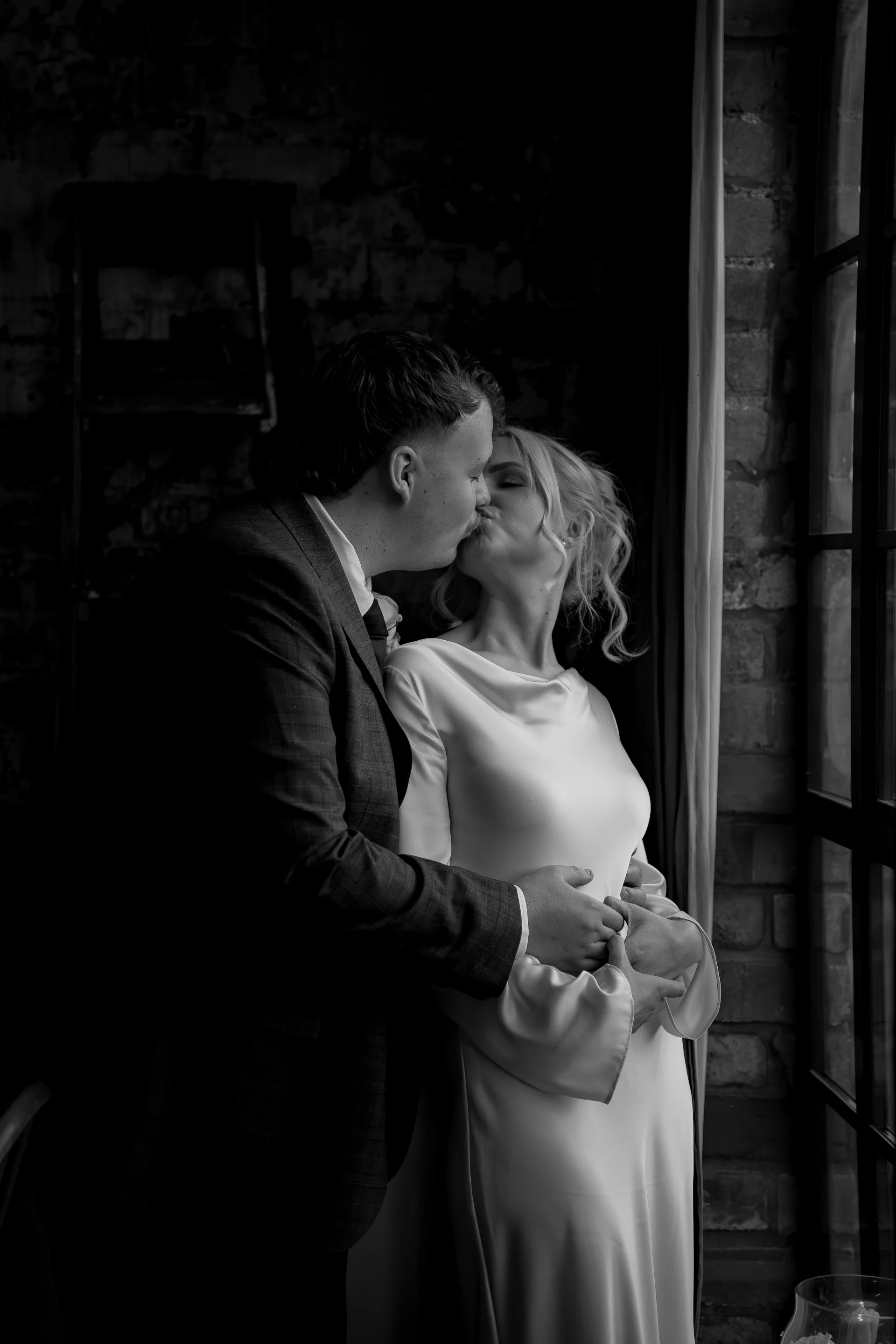 A newlywed couple shares an intimate kiss beside a large window inside an industrial venue with exposed brick.