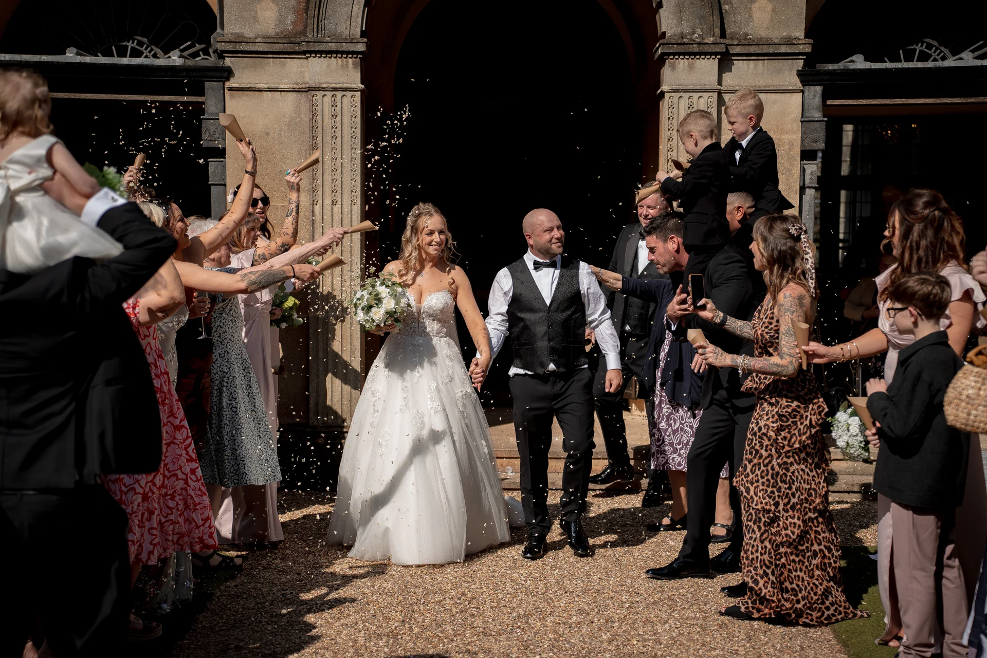 The newlyweds joyfully walk out from a grand stone archway of a country house as wedding guests shower them with a cascade of confetti.