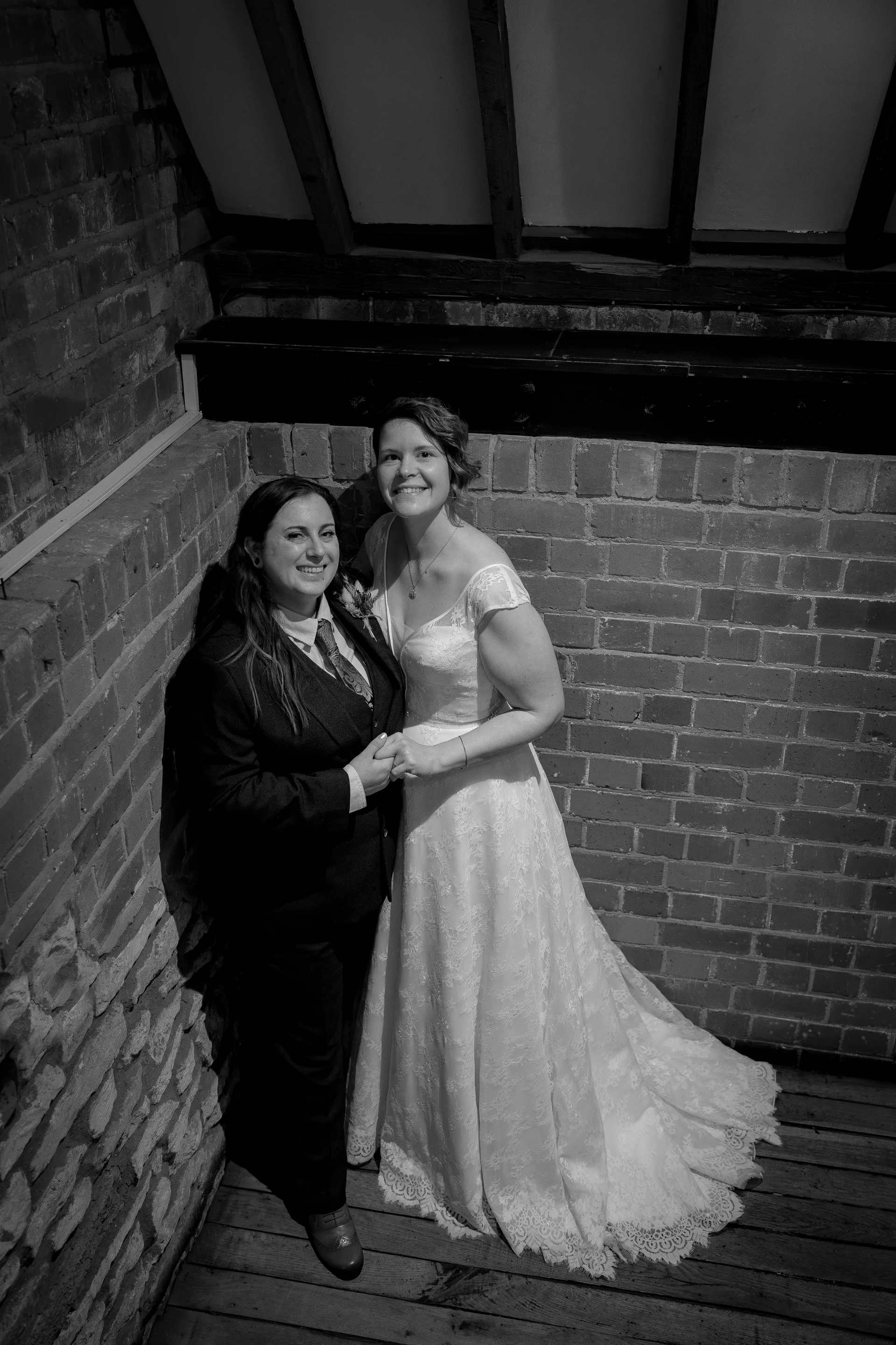 A smiling same-sex couple poses together in a black-and-white portrait against an exposed brick wall inside a rustic barn venue.
