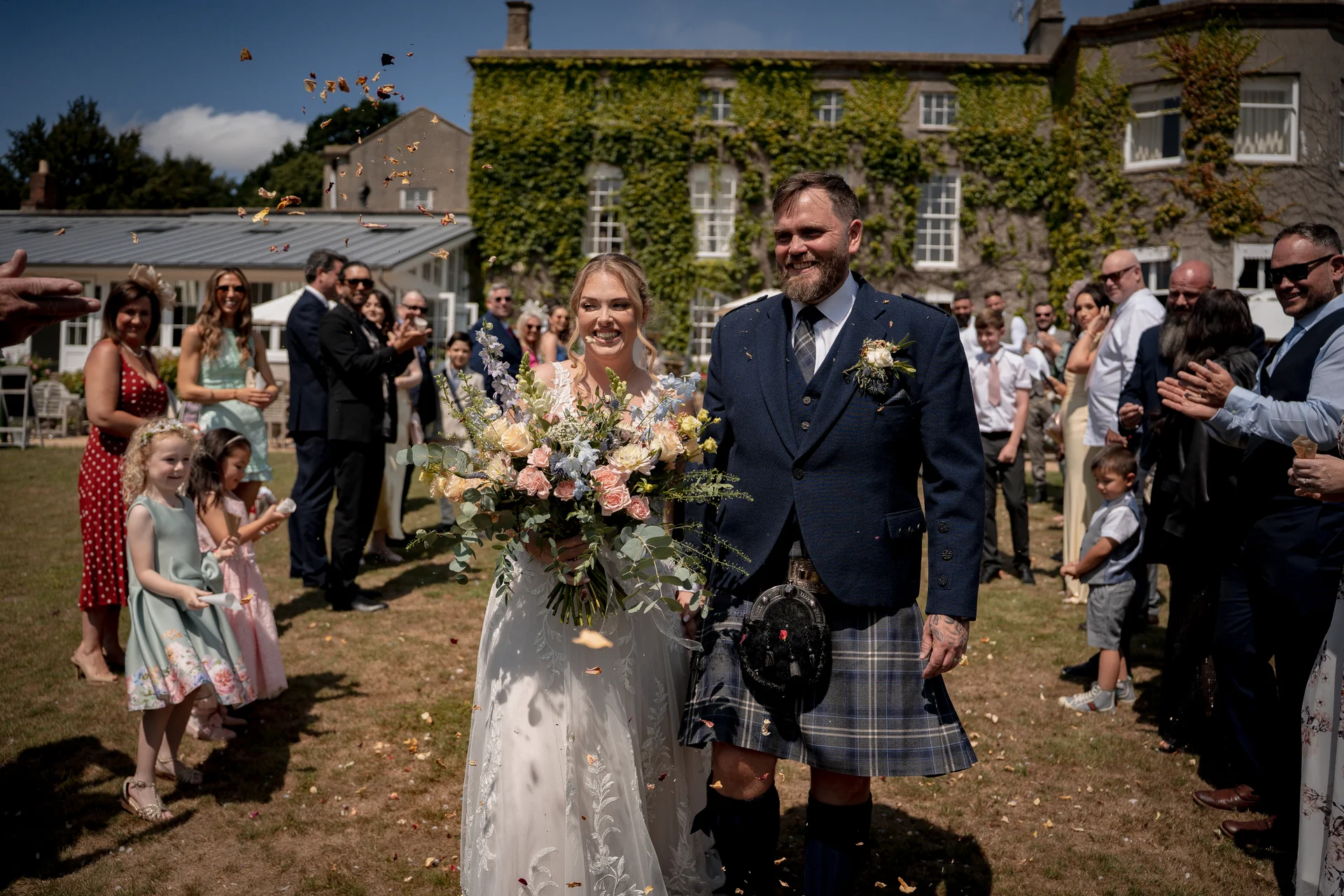 A smiling bride and groom walk hand in hand through a shower of confetti on the lawn of an ivy-clad country house while guests applaud.