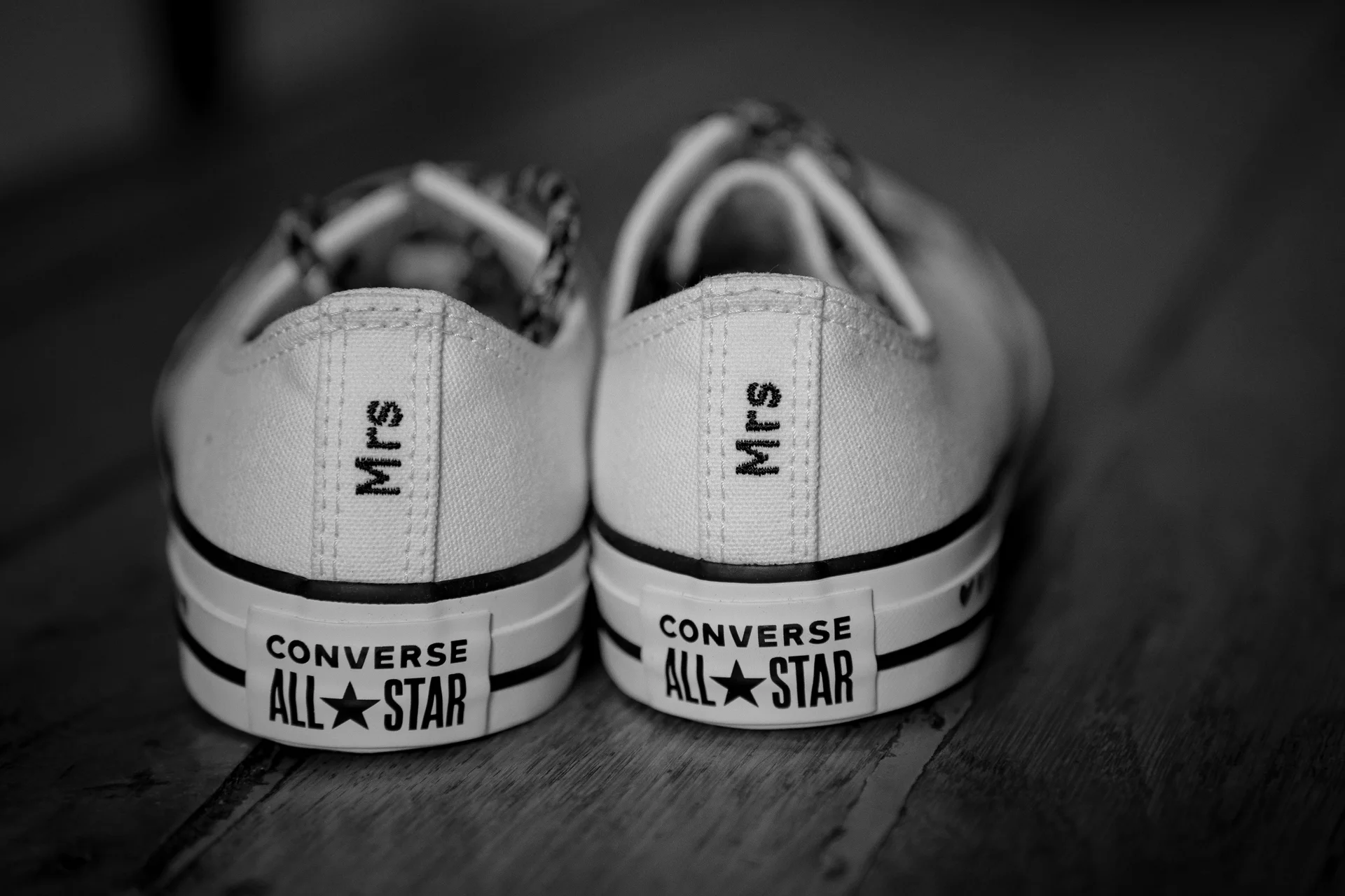 A close-up black and white photograph of two matching white Converse trainers featuring custom Mrs lettering on the back heel strips, resting on a wooden floor.