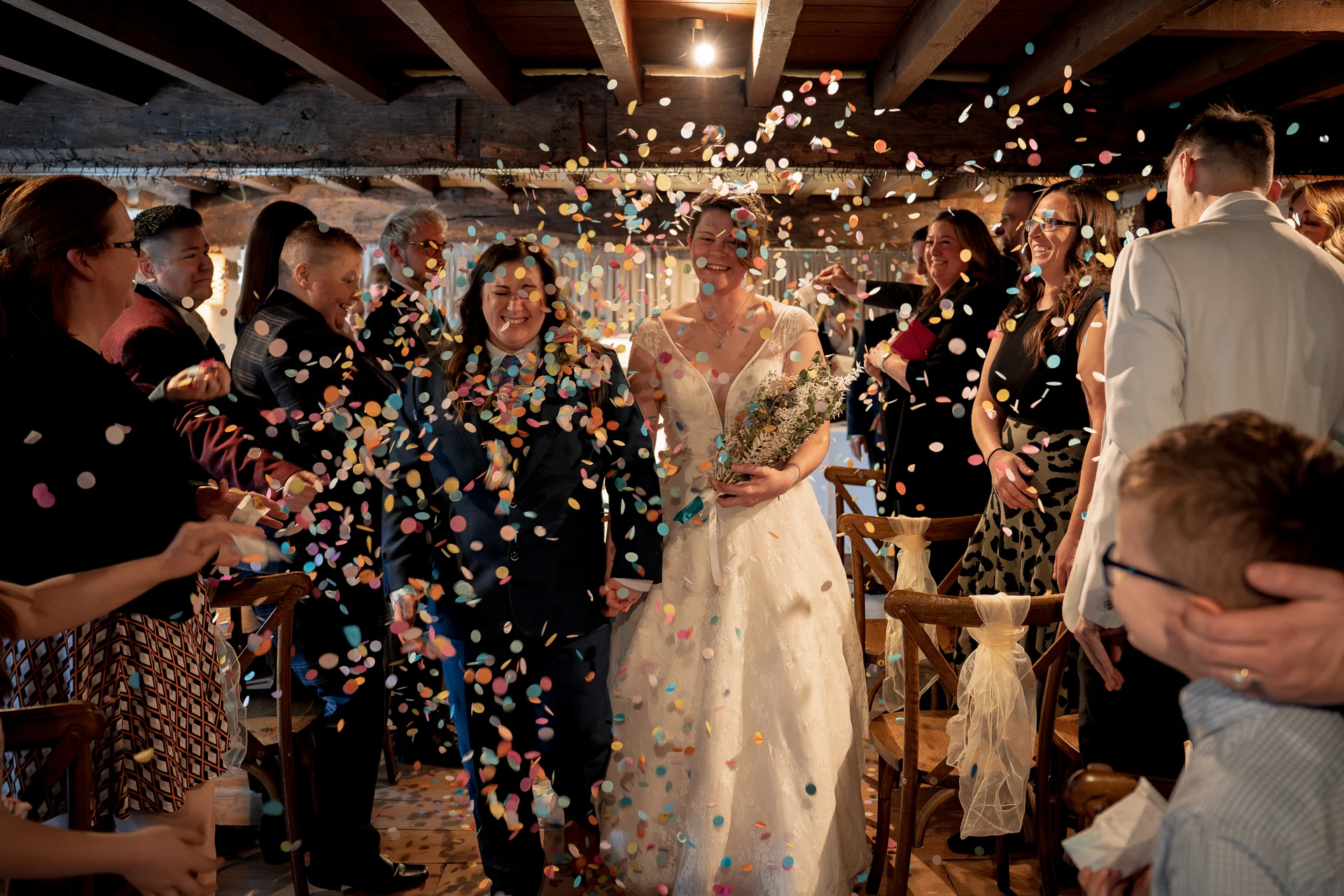 A newlywed same-sex couple walk hand-in-hand down the aisle in a rustic barn venue while cheerful guests shower them with colourful confetti.